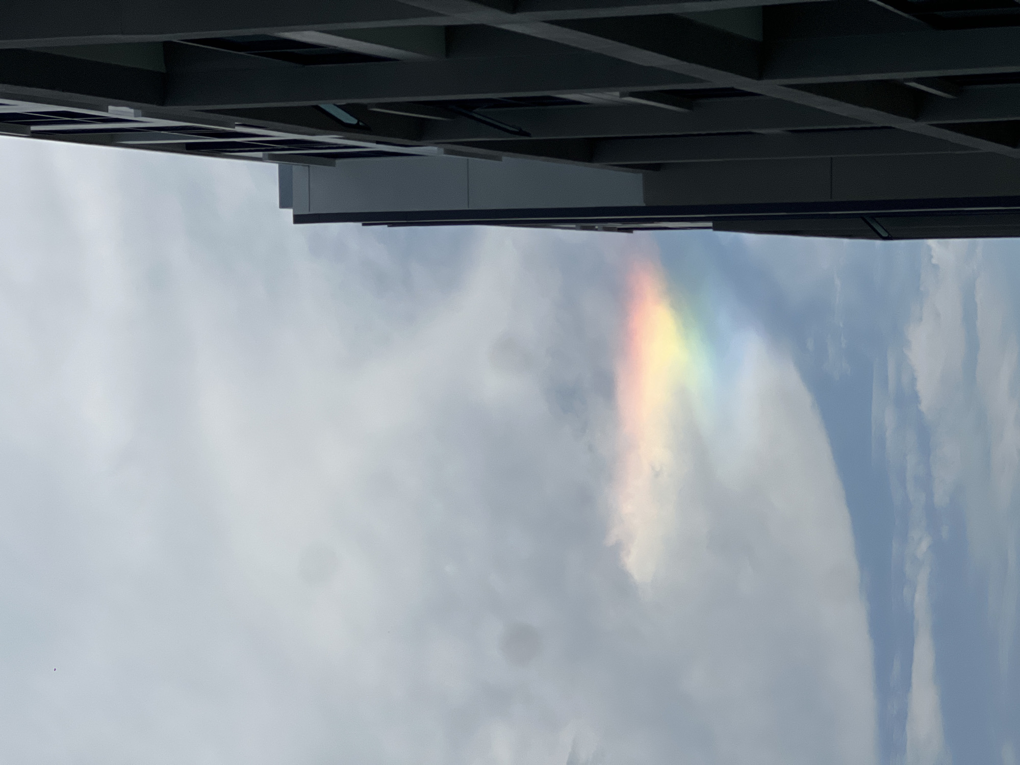 Captured from below a gray building in Choeng Thale, Thailand, a vibrant rainbow spans across a cloudy sky. The building's architectural details are visible against the bright sky. The image is rotated, presenting the building at the top of the frame and the sky below.