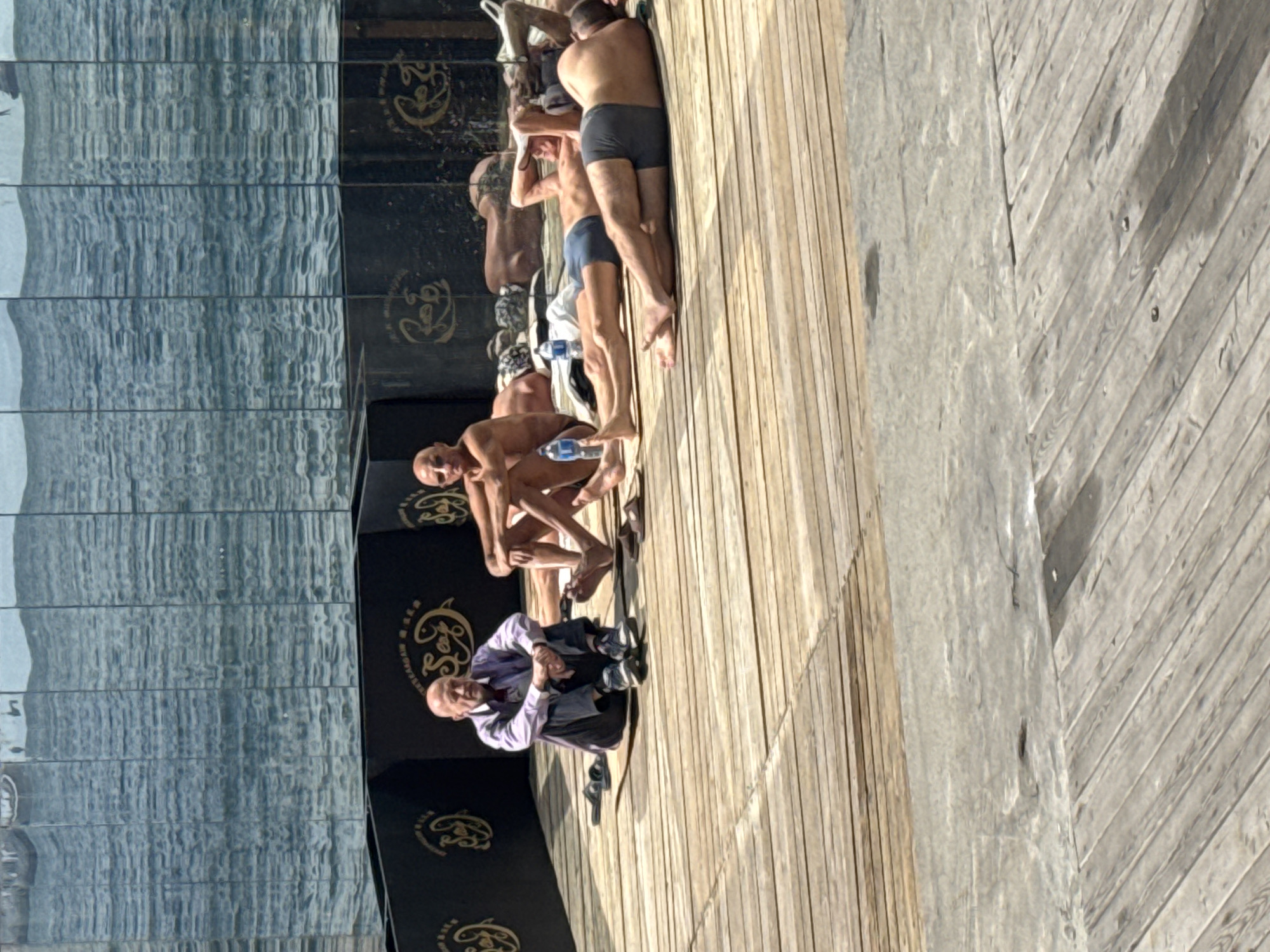 The image shows a group of people on a wooden deck or pier with a glass wall in the background, likely reflecting the sea. A man in a light-colored shirt and dark pants is sitting on the deck. A line of mostly shirtless men in swimwear are standing or crouching near the glass wall. There is a logo on the wall and water bottles near some of the men. The overall setting suggests a public beach area or recreation facility. This image was taken in Vladivostok, Russia.