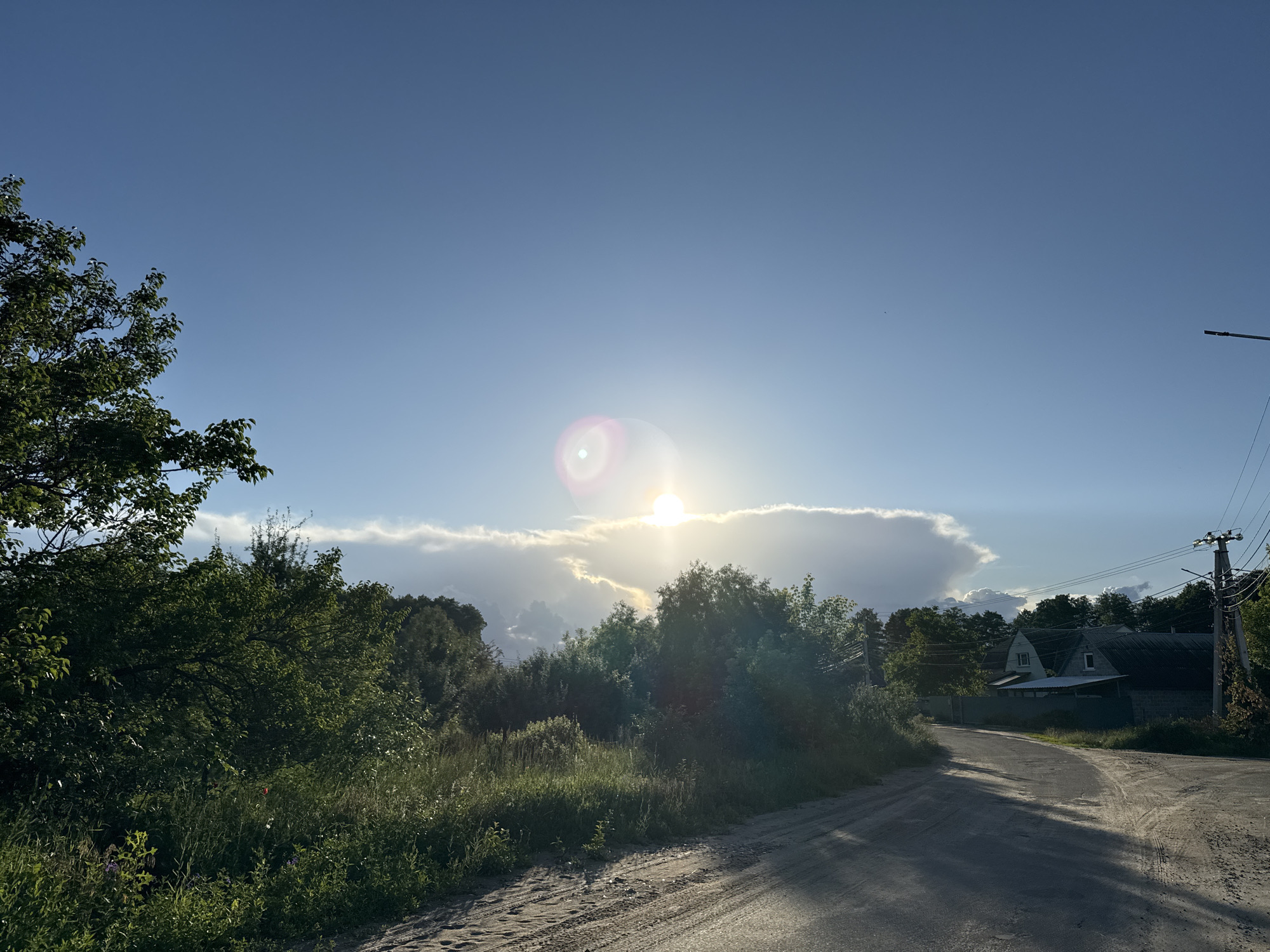 The image captures a rural landscape on a bright, sunny day. A dirt road curves through an area with abundant green foliage, including trees and tall grass. In the background, a few houses are visible amidst the trees, suggesting a small settlement. The sky is a clear blue, with the sun shining brightly and creating a lens flare. A single utility pole with wires stands on the right side of the road. The overall scene evokes a sense of tranquility and natural beauty.
