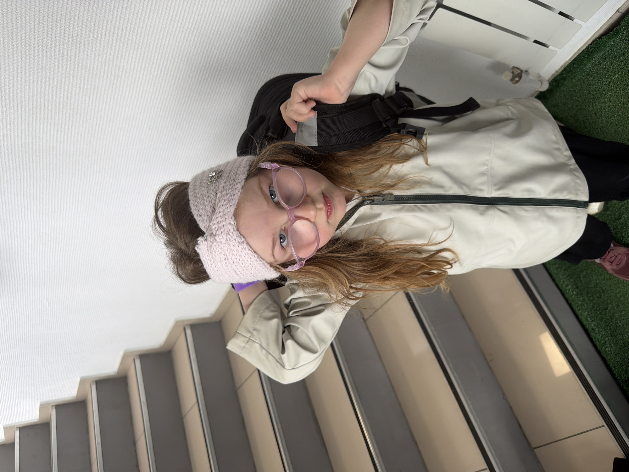 A young girl with reddish-brown hair, wearing a light pink knitted headband and large, round pink glasses, stands near a staircase and a wall, holding the straps of a black backpack. She is wearing a light-colored jacket over black pants and maroon shoes. The staircase is gray and beige, adjacent to a white wall. Beyond the wall is a patch of artificial green turf. The image was taken at an odd angle, but based on the architecture, it may be in Chaikovsky, Russia.