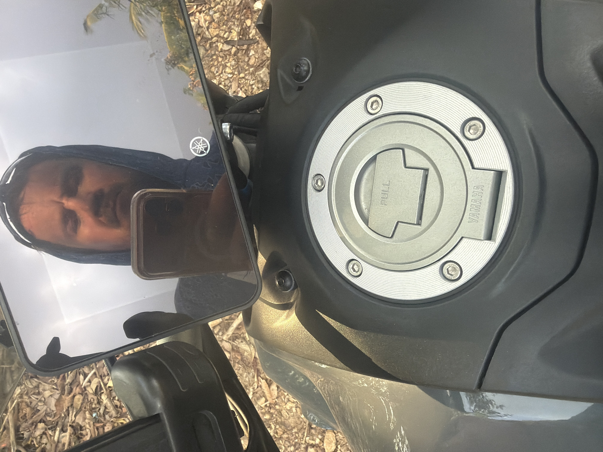 The image shows a close-up view of a motorcycle's fuel tank and a reflection in its mirror. The fuel tank cap is silver with "PULL" and "YAMAHA" text embossed on it, secured by several screws. The motorcycle is dark gray and appears to be parked outdoors, with dry leaves and soil visible in the background. In the mirror's reflection, a person wearing a hooded sweatshirt is holding a smartphone, seemingly taking the photograph. The reflection also shows foliage and a bright sky. The scene is set in Baan Thong Po, Thailand.