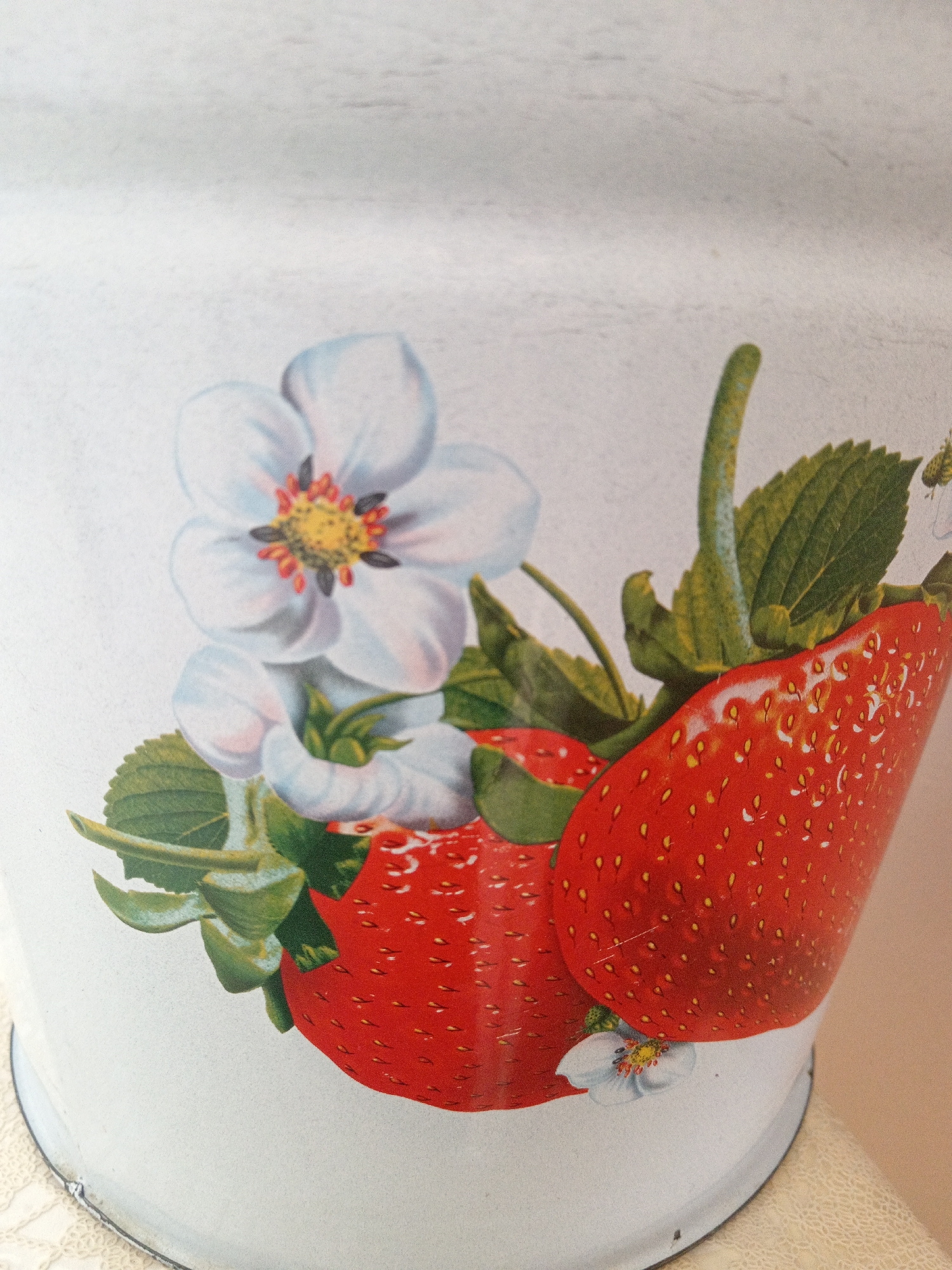 This is a close-up, top-down shot of a white, cylindrical object, possibly a container or a wall, decorated with a painted illustration of strawberries and white flowers. Two ripe, red strawberries with visible seeds and green leaves are prominently featured, along with a cluster of delicate white flowers with yellow and red centers. The illustration has a vintage aesthetic. The background is a plain, textured white surface. The lower portion of the image reveals the curved edge of the white object and a light-colored, textured surface beneath it, suggesting it is sitting on a table or shelf. There are no people or animals present. The image does not contain any information about its location in the city of Uchkeken, Russia.