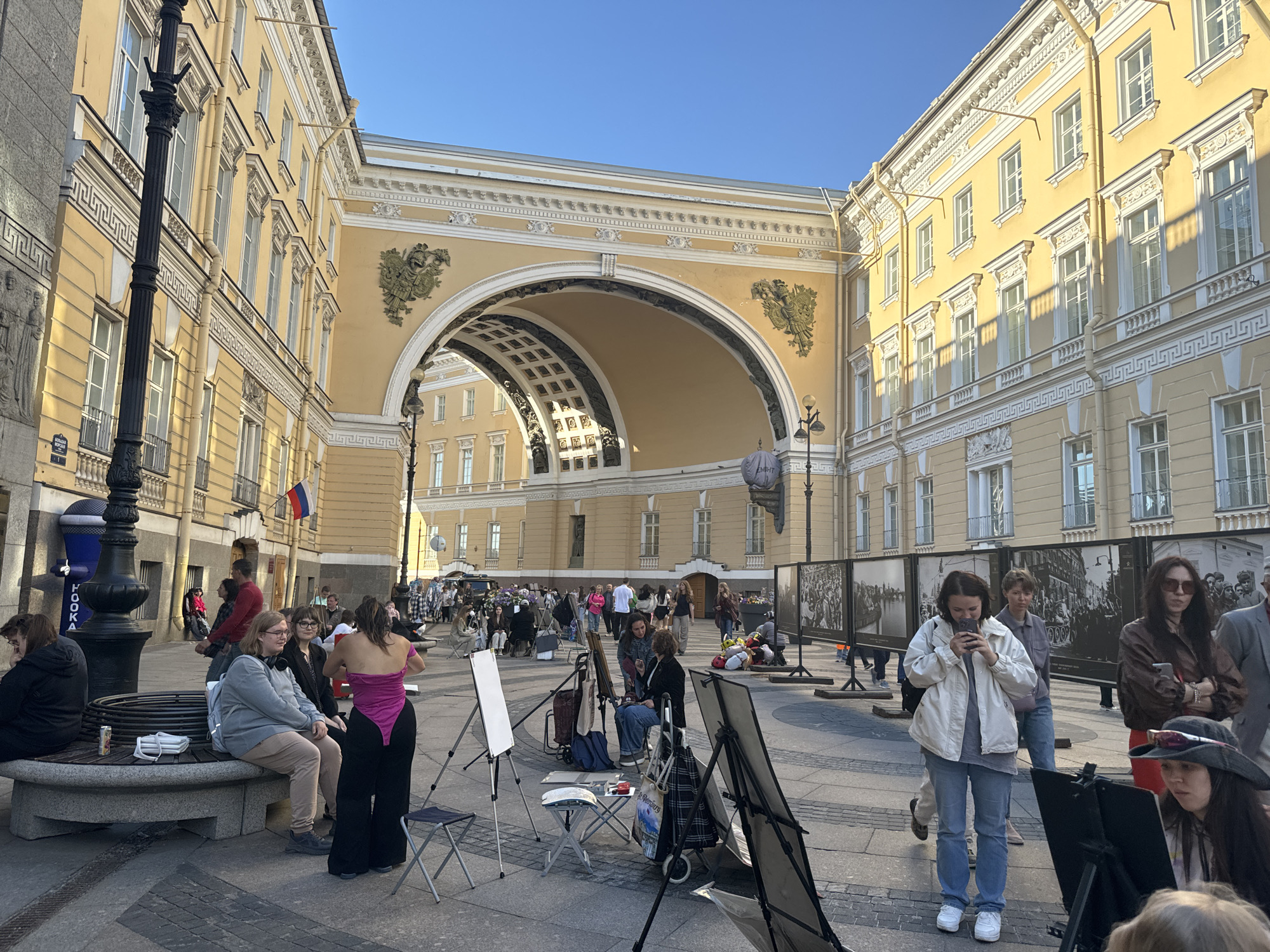 The image depicts a wide view of Palace Square in Saint Petersburg, Russia, on a bright, clear day. The iconic Triumphal Arch, part of the General Staff Building, dominates the background, its pale yellow facade adorned with intricate architectural details and gilded eagles. The sun casts long shadows across the cobblestone pavement, indicating it is likely late afternoon.
In the foreground, several people are engaged in various activities. A group of individuals appears to be participating in an outdoor art session. Easels with blank canvases are set up, and some people are seated, possibly sketching or painting. Among them, a woman in a vibrant pink leotard and black wide-legged pants stands out. Another woman, wearing a white jacket and jeans, is looking at her phone while standing.
To the left, a large, ornate lamppost stands near a curved stone bench where several people are resting. A blue cylindrical structure with the word "HOOОKA" on it is visible behind the lamppost. Along the right side of the square, large black and white photographic displays are mounted on stands, showcasing historical scenes.
The overall atmosphere is one of civic life and public gathering, with a mix of tourists and locals enjoying the expansive urban space and architecture. The clear blue sky and warm sunlight contribute to a pleasant, inviting scene.