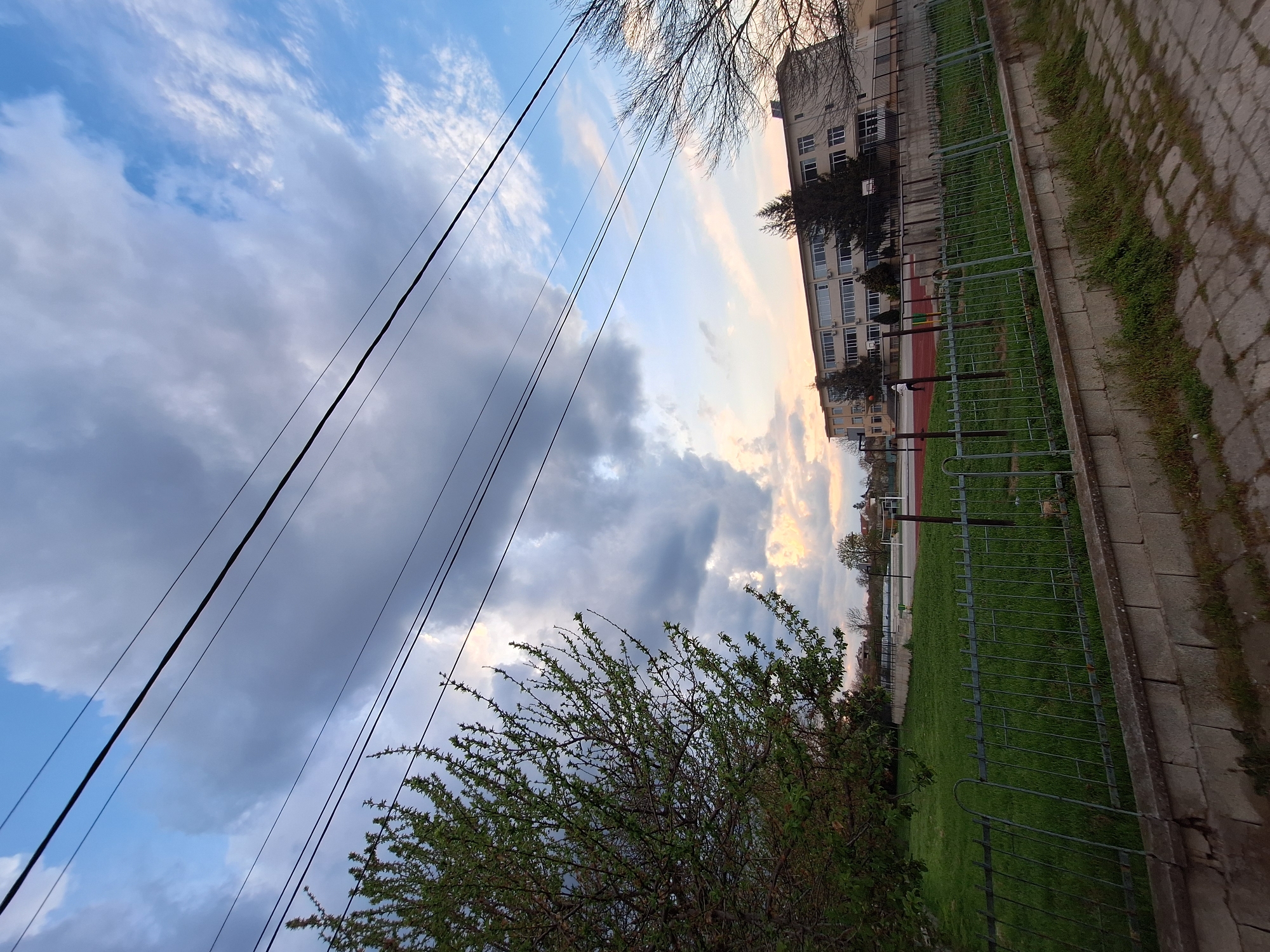 This image captures an outdoor scene in Dolni Chiflik, Bulgaria, depicting a mix of natural elements and urban structures, likely during late afternoon or early morning due to the lighting.
The sky dominates the upper portion of the frame, displaying a dynamic mix of clear blue patches, fluffy white clouds, and darker grey clouds. Several dark utility cables or power lines stretch diagonally across the sky, adding a linear element to the composition.
In the mid-ground, a multi-story building, possibly an apartment complex or a school, is visible with numerous windows. In front of the building lies a green field, which appears to be a sports ground given the visible red track surface and what looks like a basketball hoop structure. A metal fence separates this green area from a paved walkway or path made of concrete slabs. Grass and weeds grow along the edges of the path and between the slabs.
The foreground on the left features a vibrant green bush or small tree with new foliage, indicating spring or early summer. Another tree with bare branches is visible towards the top right, near the building.
There are no people or discernible activities taking place, presenting a tranquil, static view of the environment. No text is visible within the image.