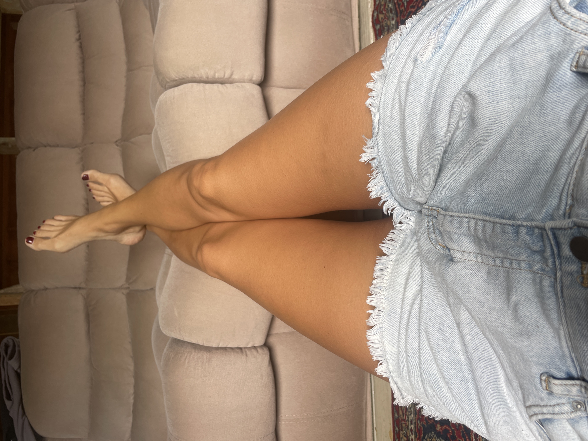 A high-angle, full shot depicts a woman's legs crossed on a beige sofa. She is wearing light blue, denim shorts with a frayed hem. Her toenails are painted a dark red color. The background includes a portion of a dark patterned rug. There is no indication of the location being Baranovka, Russia.