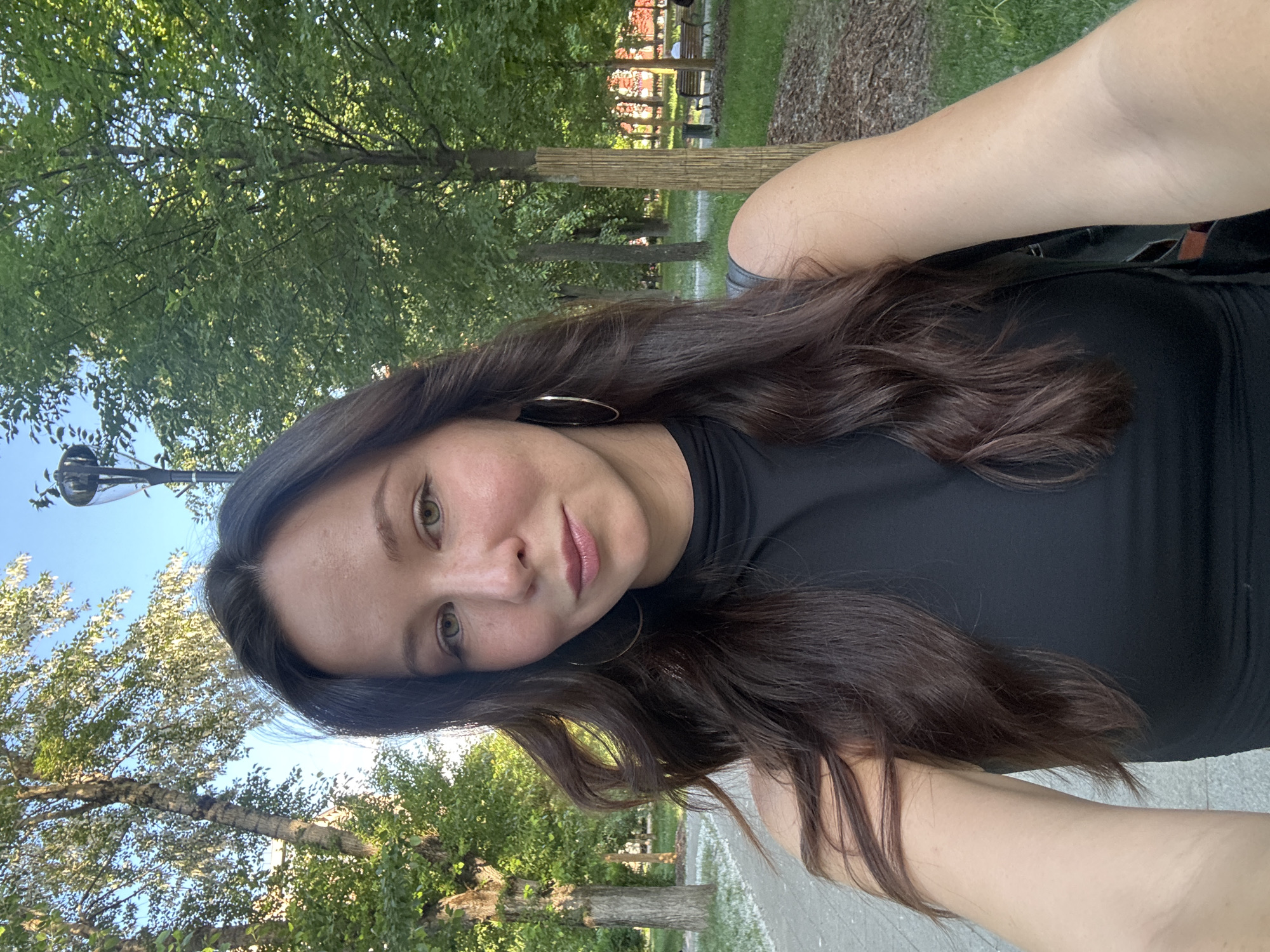 The image is a selfie of a young to middle-aged woman taken outdoors in a park or green space, likely in Moscow, Russia. The woman, positioned in the foreground, has long, wavy dark brown hair and light green or hazel eyes. She is looking directly at the camera with a gentle, pleasant expression and is wearing a black, high-necked top along with large golden hoop earrings. Her arms are visible, suggesting she is holding the phone to take the photograph.
The background is filled with lush green trees and foliage under a bright blue sky, indicating clear, sunny weather during a warm season such as late spring or summer. Sunlight filters through the tree canopy, illuminating the scene. In the mid-ground, a paved or dirt path is visible, and further back, elements like park benches and a structure with red accents can be seen, consistent with a public park environment. A light pole or similar outdoor fixture is partially visible on the left side. The overall mood of the image is calm and natural. No visible text is present.