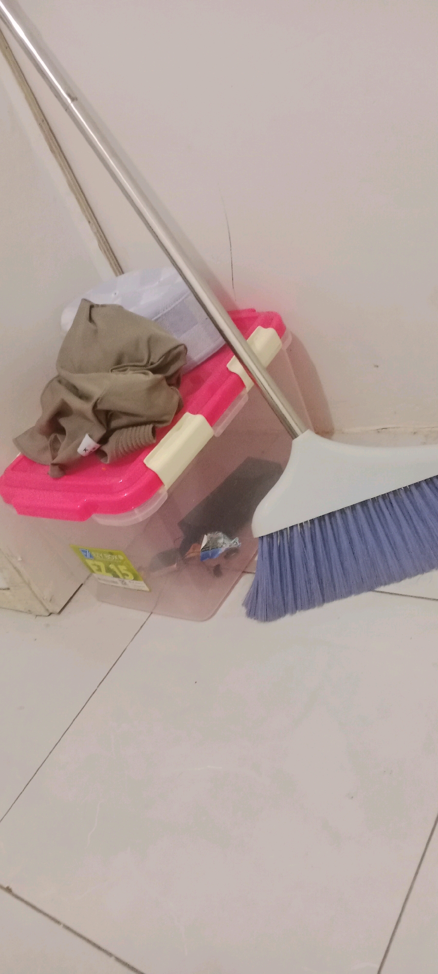 A metal broom with a purple brush rests against a light-colored wall in what appears to be a domestic storage area. The broom is positioned next to a translucent pink and clear plastic container, which holds a white cloth, a folded tan garment, and what seems to be a small, crumpled piece of shiny material. The container is on a light-tiled floor. The scene is illuminated by artificial light, casting soft shadows. The image does not depict any people or actions beyond the placement of objects. The overall setting suggests a utilitarian space, possibly a utility closet or a corner of a room where cleaning supplies are kept. The location is identified as Serongga, Indonesia.