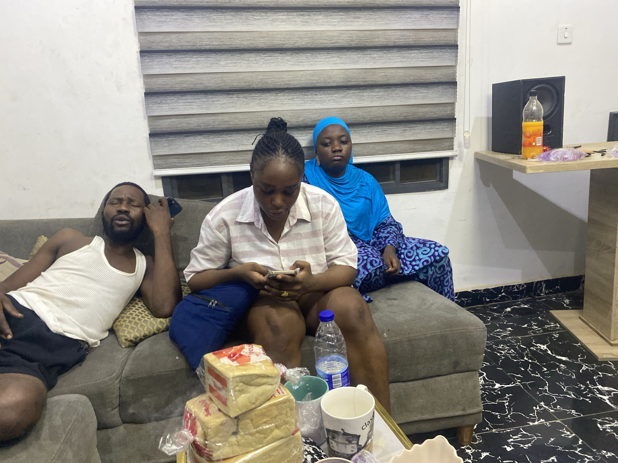 Three individuals relax in a casually furnished indoor living space, likely a living room in Abuja, Nigeria. On the left, a man with a beard, wearing a white tank top and black shorts, reclines on a grey sofa. He holds a phone to his ear, with his eyes closed or nearly closed, appearing to be resting or listening intently. In the center, a woman in a light-colored striped button-up shirt and blue shorts sits upright, engrossed in a smartphone held in both hands. Her hair is pulled back, and she wears a ring on her right ring finger. Behind her, a woman in a bright blue hijab and a patterned blue dress sits, looking towards the viewer with a neutral, slightly pensive expression.
In front of the sofa, a low coffee table holds several items, including stacked loaves of bread wrapped in plastic, with "FARMHOUSE" and "BUTTERBREAD" partially visible on the packaging. Also on the table are a clear plastic water bottle with a blue cap and several mugs, one white mug showing the partial text "clas". To the right, a black speaker and a bottle of orange juice, possibly "PULPY ORANGE," sit on a dark wooden table, alongside small clear plastic bags containing purple items. The room features horizontal striped window blinds and black and white marble-patterned floor tiles. The diffused lighting suggests a relaxed atmosphere during either daytime with blinds drawn or evening.