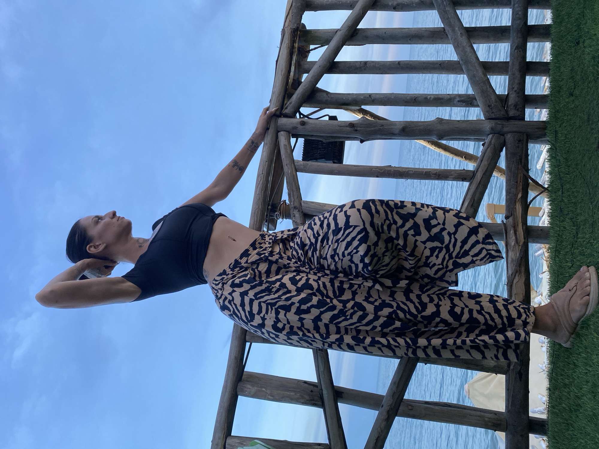 A full-body shot captures a woman standing on a grassy area next to a wooden railing. She is facing to the right, looking out at the ocean. Her left arm is raised, with her hand behind her head, and her right hand is resting on the railing. She is wearing a black crop top and a flowing skirt with a tan and black zebra-like pattern. The ocean is visible in the background, with a blue sky overhead. The location is identified as Phu Thuy Ward, Vietnam.