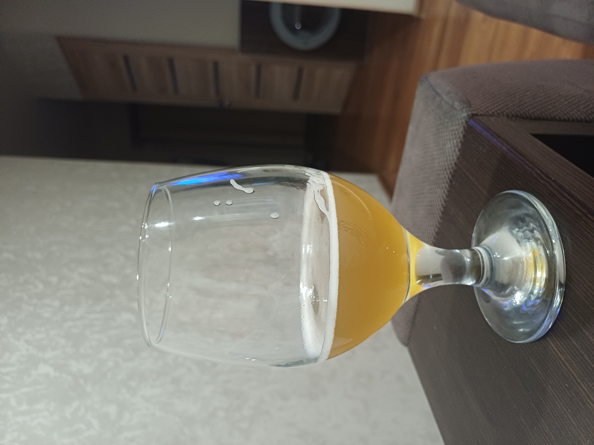 A partially consumed glass of beer sits on a dark brown wooden table. The glass is clear and stemmed, with a wide bowl. The beer is a light amber color and has a thin layer of white foam on top. The table appears to be part of a piece of furniture, possibly a coffee table or side table, near an upholstered surface. A cabinet is visible in the background. The location is Chaikovsky, Russia.