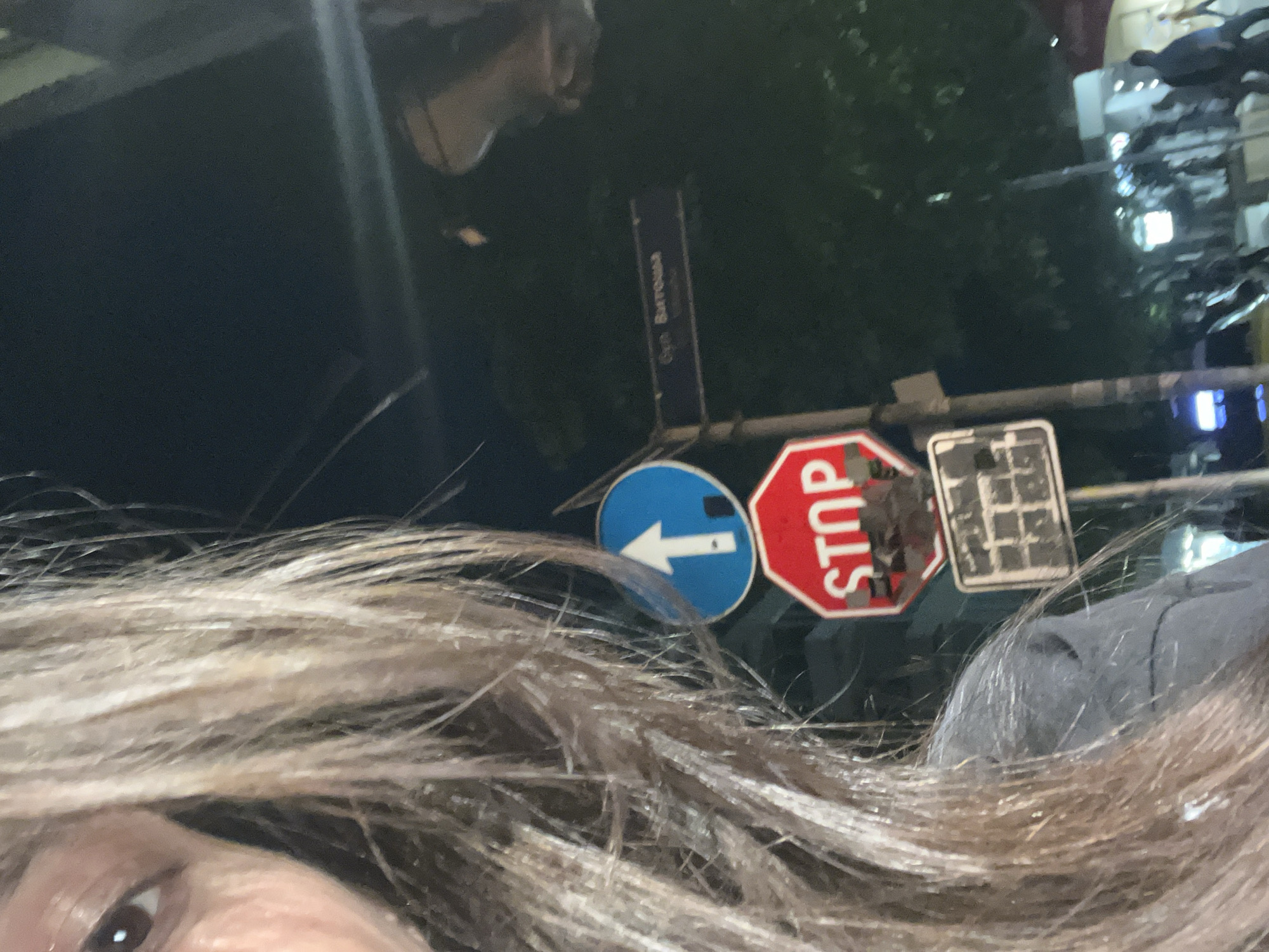 The image is a close-up shot of a person's eye and hair, with a street scene in the background. The person's eye is brown and appears to be looking to the left. Their hair is long, brown, and slightly wavy. In the background, there are several street signs, including a blue circular sign with a white arrow pointing upwards and a red octagonal "STOP" sign. There is also a street name sign with Cyrillic text that reads "ул. Витоша" (ul. Vitosha). The environment appears to be a city street at night, with streetlights and some blurred figures of people in the distance. The presence of the "Vitosha" street sign indicates that the location is likely on Vitosha Boulevard in Sofia, Bulgaria, a well-known commercial street. The scene suggests a casual moment captured by the person, possibly while walking or standing on the street.
