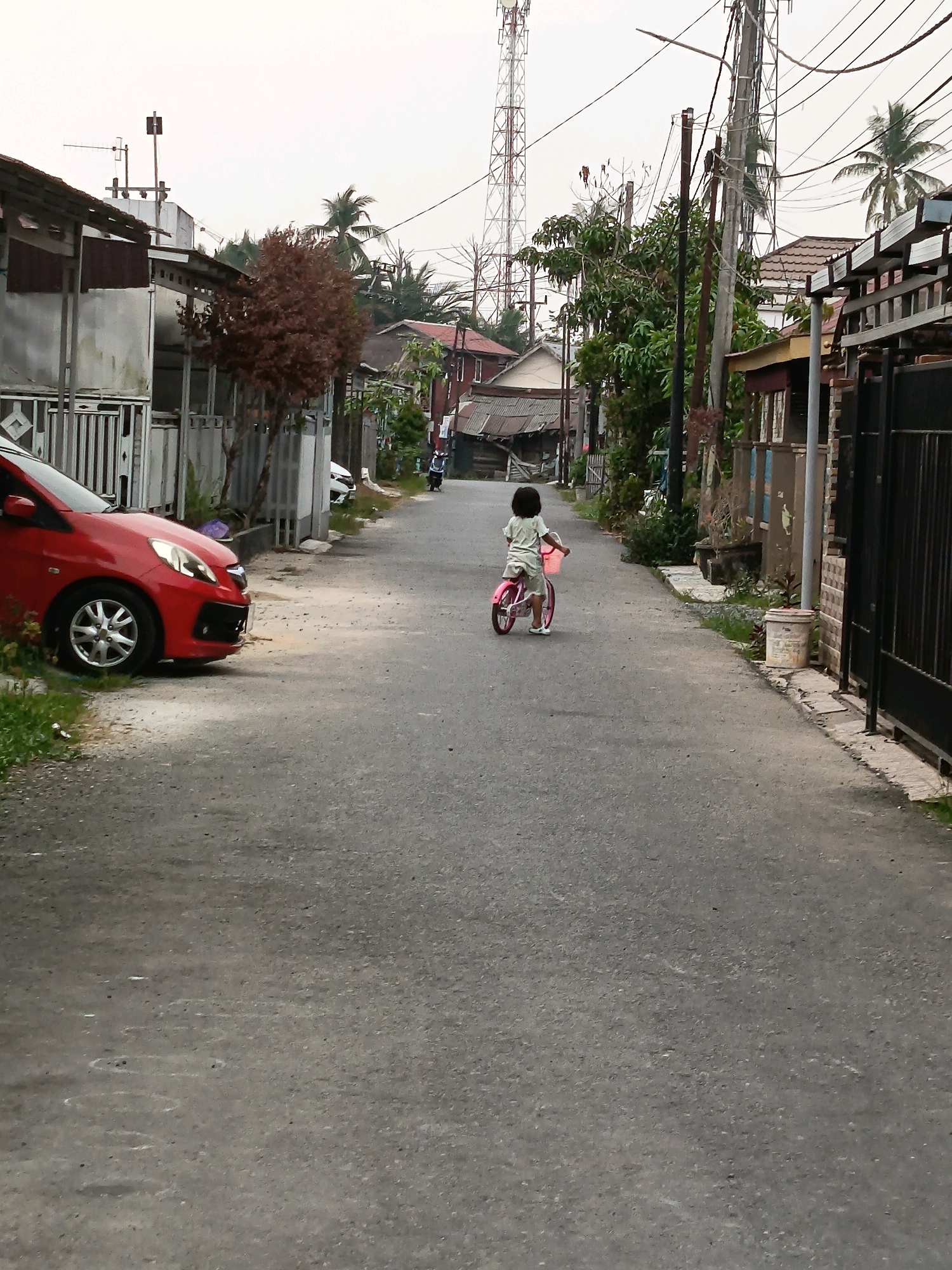 A young child, viewed from behind, rides a small pink bicycle down a paved street in Banjarmasin, Indonesia. The child wears a light-colored t-shirt and shorts. The street is lined with houses and some vegetation, including palm trees. A red car is parked on the left side of the street. In the distance, a tall tower is visible against an overcast sky.
