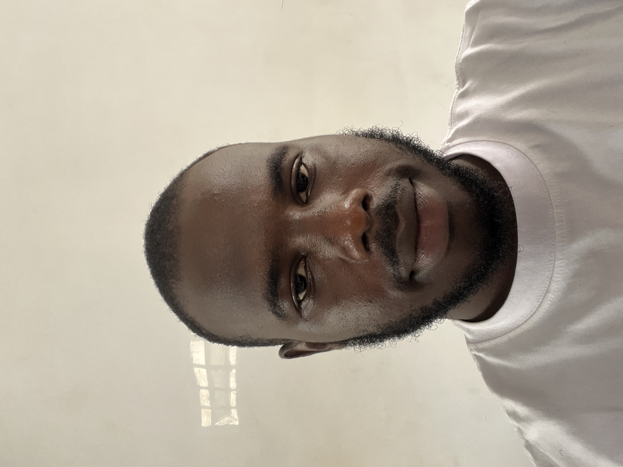 A close-up studio shot depicts a man of African descent with dark brown skin and a closely shaved head. He has a dark, well-groomed beard. The man's expression is neutral, with his eyes focused on the camera. He is wearing a plain white t-shirt. The background appears to be a plain, off-white or light-colored wall. There is no indication of the location being in Jos, Nigeria, or any other city. There are no visible subjects, objects, actions, or interactions present in the image besides the individual. The environment is a plain studio setting with minimal distractions.