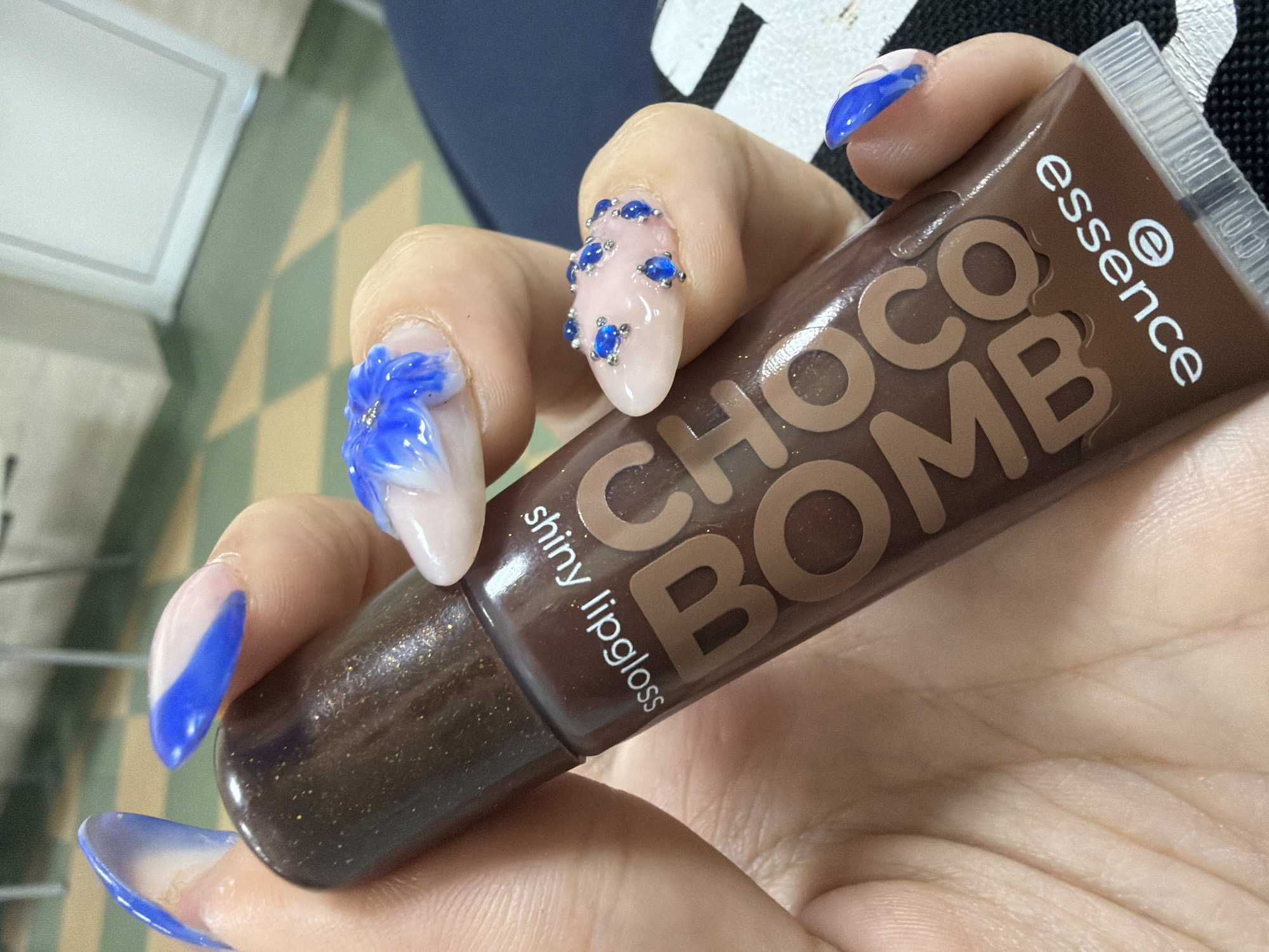 A close-up shot focuses on a person's hand holding a tube of "essence Choco Bomb shiny lipgloss." The lipgloss tube is brown with the brand name and product name printed in a lighter brown font. The tube itself has a shimmery, textured appearance. The person's fingernails are elaborately decorated with light pink polish and detailed blue designs, including what appear to be blue flowers and blue rhinestones on some nails. The hand is positioned over a surface with a checkered pattern of green and cream squares, possibly a floor or table. The background is out of focus, but shows some hints of furniture or room elements. The lighting suggests an indoor setting, possibly during the daytime. The image conveys a sense of personal care and aesthetic presentation. There is no indication of the specific time of day, weather, or emotional state of the person. The visible text clearly identifies the product as "essence Choco Bomb shiny lipgloss."
