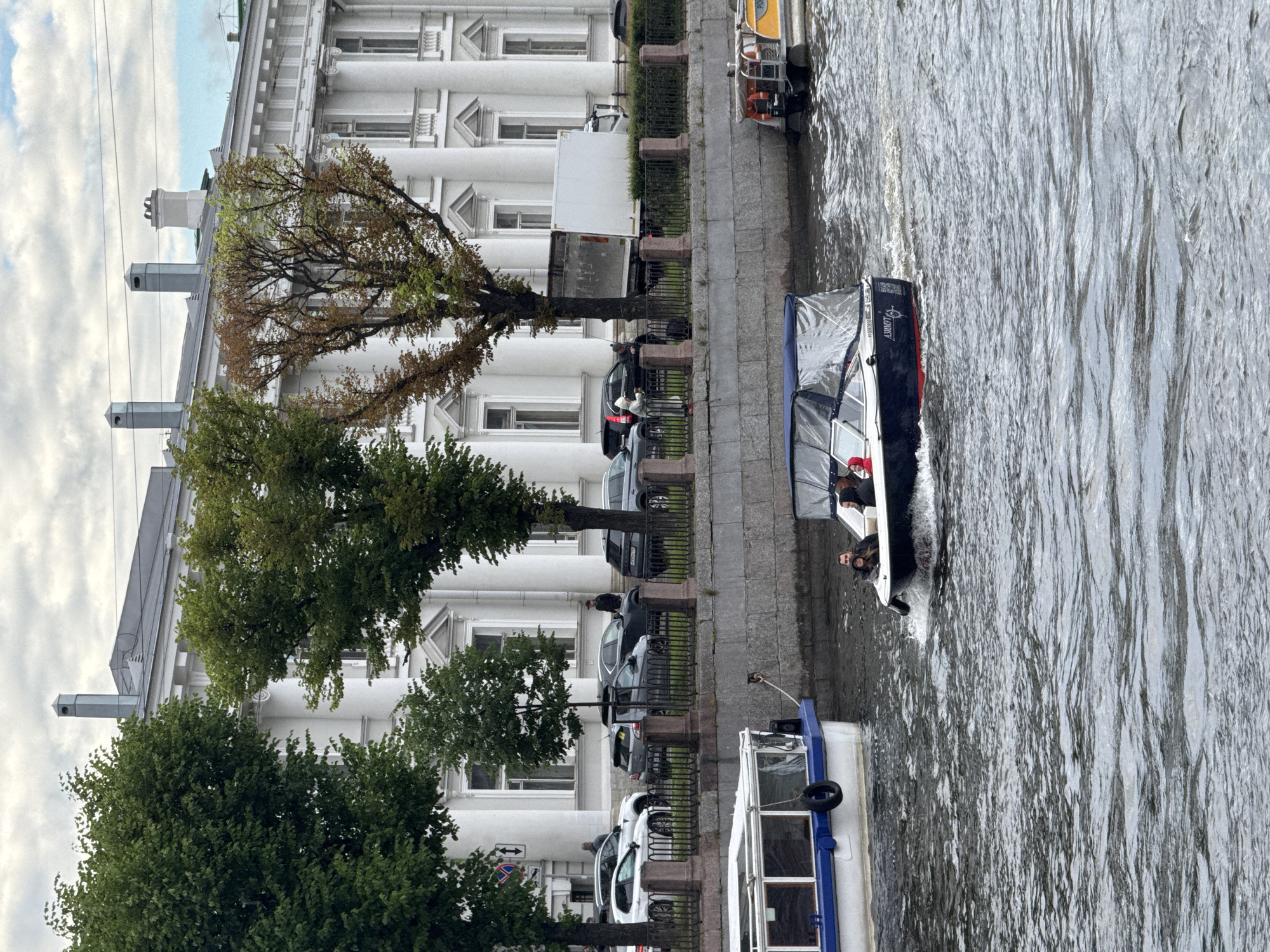 The image shows a canal in Saint Petersburg, Russia, with a white, classical-style building on the left and a waterway on the right. Several boats are visible on the canal, including a dark blue tourist boat with a canopy and people on board, and a white boat with blue trim. Cars are parked along the embankment next to the building, and trees with green and brown foliage are scattered along the canal. The sky is overcast. The image appears to have been taken during the daytime. The building features columns and multiple windows, indicative of historical architecture. The boats are moving through the water, suggesting they are part of a tour or transport service. There is some visible text on the dark blue boat, which appears to be "LUVY".