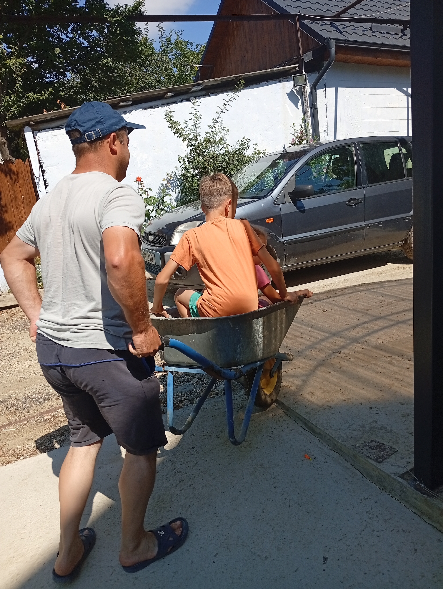 In Pănășești, Moldova, a man wearing a grey t-shirt, dark shorts, a blue baseball cap, and blue slides pushes a wheelbarrow containing two children. The boy in front, wearing an orange t-shirt and green shorts, sits facing forward, while a younger child in a pink shirt is partially visible behind him. The wheelbarrow has a blue frame and a yellow wheel. Behind them, a dark grey car is parked next to a white building with a dark grey roof. The scene is illuminated by bright sunlight, casting shadows on the concrete ground.