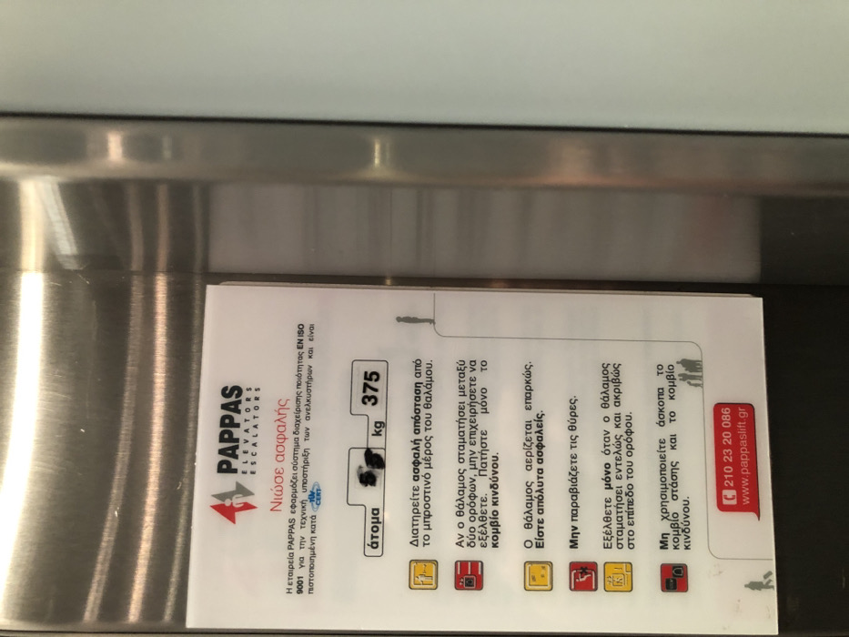 The image displays a white safety information sticker or document affixed to a brushed or polished metallic surface, likely inside an elevator cabin. The document is oriented vertically, with its text in Greek, and is partially obscured by reflections on the metal.
The sticker provides information from "PAPPAS ELEVATORS ESCALATORS" under the heading "Νιώσε ασφαλής" (Feel safe). It states that the PAPPAS company implements an EN ISO 9001 quality management system for elevator technical support and is TÜV CERT certified. Contact details include a phone number, 210 23 20 086, and a website, www.pappaslift.gr.
Safety instructions are presented with accompanying pictograms:
- The elevator capacity is stated as 375 kg, with a partially visible number of "άτομα" (persons).
- Users are advised to "Διατηρείτε ασφαλή απόσταση από το μπροστινό μέρος του θαλάμου" (Maintain a safe distance from the front of the cabin).
- In case the cabin stops between floors, users should "μην επιχειρήσετε να εξέλθετε. Πατήστε μόνο το κομβίο κινδύνου" (not attempt to exit, but only press the emergency button).
- An assurance is given that "Ο θάλαμος αερίζεται επαρκώς. Είστε απόλυτα ασφαλείς" (The cabin is adequately ventilated. You are completely safe).
- A warning states: "Μην παραβιάζετε τις θύρες" (Do not force the doors).
- Users should "Εξέλθετε μόνο όταν ο θάλαμος σταματήσει εντελώς και ακριβώς στο επίπεδο του ορόφου" (Exit only when the cabin has stopped completely and precisely at the floor level).
- Finally, it advises: "Μη χρησιμοποιείτε άσκοπα το κομβίο στάσης και το κομβίο κινδύνου" (Do not needlessly use the stop button and the emergency button).
The metallic surface reflects ambient light, creating bright streaks. The setting is indoors, with no people visible. The context suggests the image was taken in Moschato, Greece.