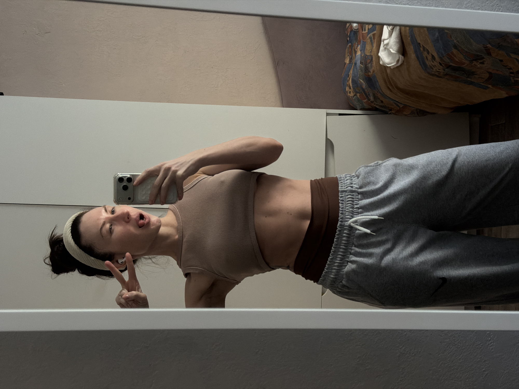 A young woman is captured in a playful mirror selfie, taken indoors in Perm, Russia. The image is oriented sideways, with the woman standing facing left, looking at her reflection. She is wearing a light brown ribbed crop top over a darker brown sports bra, paired with grey sweatpants that feature a white drawstring and elasticated cuffs. A small black Nike swoosh logo is visible on the left leg of the sweatpants. Her dark hair is pulled back with a light-colored headband, and she has white wireless earbuds in her left ear.
She holds a silver smartphone with a clear case in her right hand, taking the photo. With a cheeky expression, she sticks her tongue out slightly and forms a peace sign with her left hand. Her pose highlights her toned abdomen. The mirror reflects a room with light beige textured walls. In the background reflection, a colorful, patterned fabric item, likely a bed or sofa with a throw, is visible, along with a folded white cloth. The mirror itself appears to be a full-length one, possibly part of a white wardrobe or closet. The overall mood is casual and lighthearted.