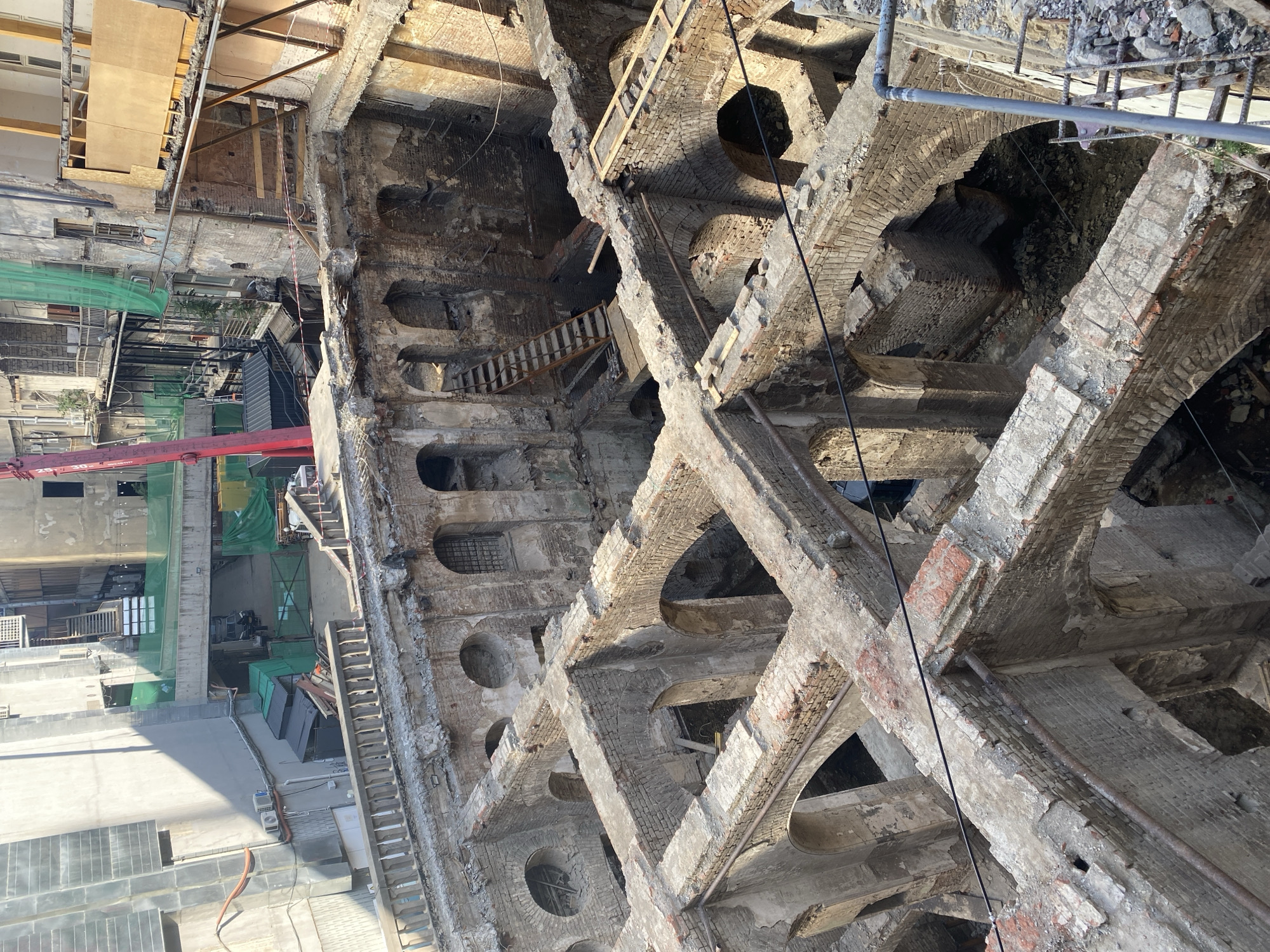 This is an aerial view of the ongoing demolition and reconstruction of the historic Gudiashvili Bathhouse in Tbilisi, Georgia. The scene depicts the exposed brickwork and arched structures of the bathhouse's interior, revealing its historical architecture. Construction materials, scaffolding, and remnants of the building's former state are visible throughout the site. In the background, adjacent buildings and construction equipment, including a red crane, indicate the urban setting of the reconstruction project. The sunlight casts shadows across the ruins, highlighting the textures of the weathered brick and stone.