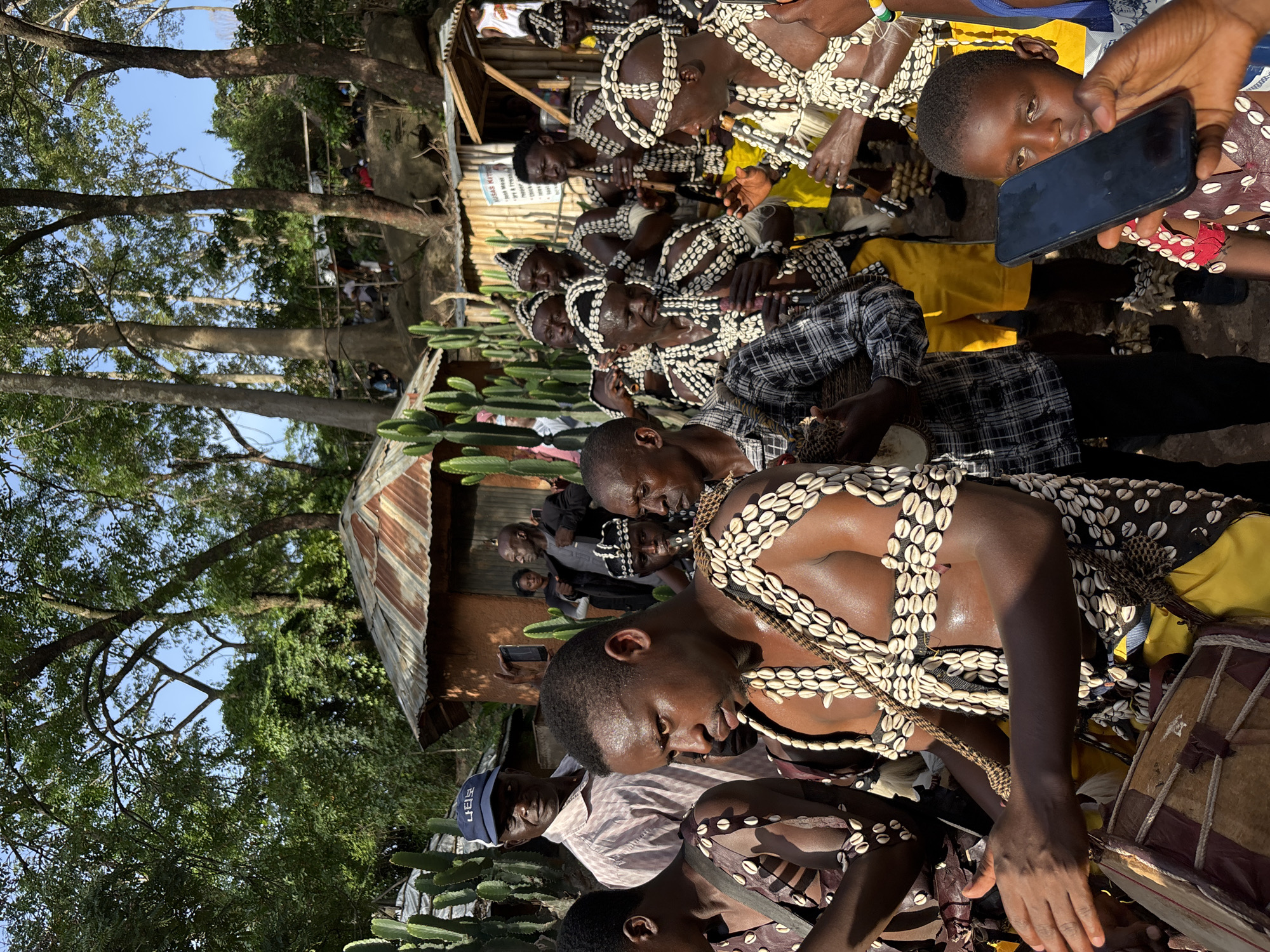 This media file captures a group of people in what appears to be a cultural event in Jos, Nigeria. The subjects are primarily young men dressed in traditional attire adorned with shell necklaces and headdresses. Some are actively playing drums, suggesting a performance or ritual. The scene is set outdoors, with a backdrop of trees, a simple building with a corrugated metal roof, and what seem to be cacti. The lighting indicates it's daytime, with sunlight filtering through the trees. The atmosphere seems to be one of active participation and perhaps celebration. One individual in the foreground is holding up a smartphone, possibly recording or photographing the event. There is a sign with text that is partially obscured and difficult to read in its entirety, but it appears to be in English and possibly another language. The overall impression is a vibrant cultural gathering in a rural or semi-rural setting.