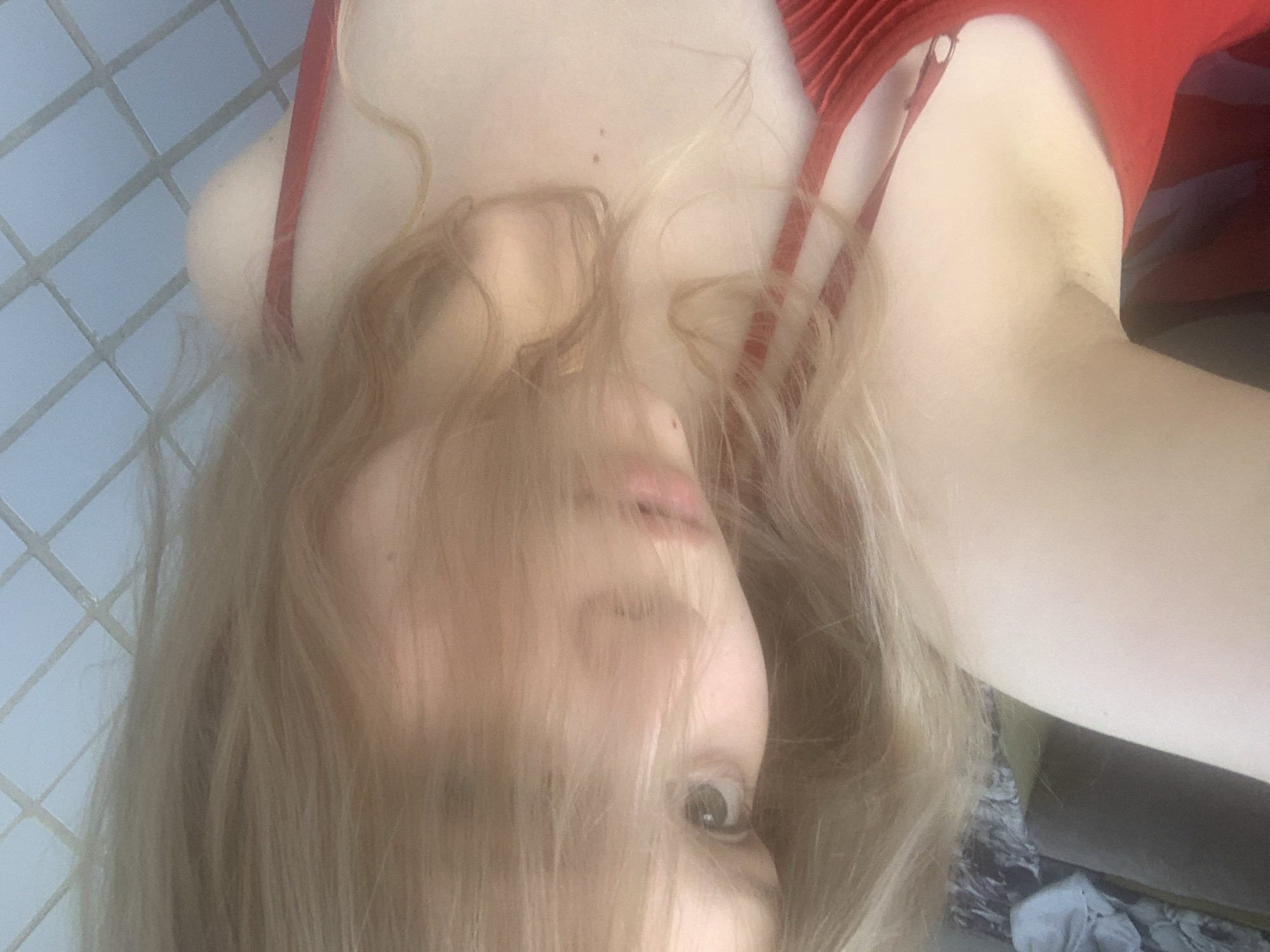 The image shows a close-up, tilted selfie of a person with blonde hair. The person is wearing a red garment, possibly a swimsuit or a top. Their face is partially obscured by their hair, but one eye and part of their mouth are visible. The background appears to be a tiled wall. There are no discernible elements that would indicate a specific location in the city of Unknown, Russia.