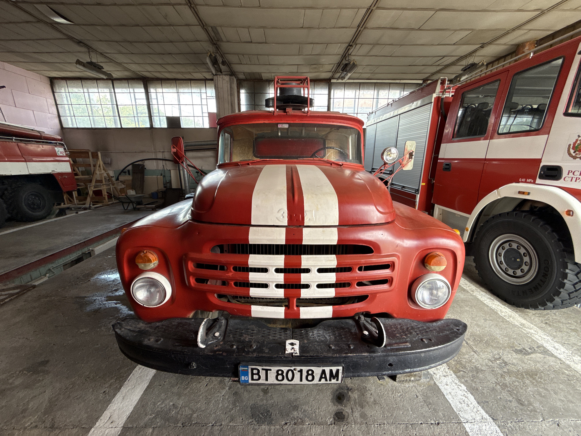 The image depicts two red and white fire trucks parked inside a fire station in Strazhitsa, Bulgaria. The foreground features the front of a vintage fire truck, characterized by a rounded cab, a white stripe down the hood, and a black bumper with a Bulgarian license plate that reads "BT 8018 AM". Behind it and to the right is a more modern fire truck, with a visible tire labeled "Continental". The left side of the image shows another fire truck that is only partially visible. The setting is a garage or fire station with overhead fluorescent lighting and a row of windows across the back wall. The floor is concrete with white lane markings. Other objects present in the background include wooden step ladders, tools and equipment, and other miscellaneous objects.