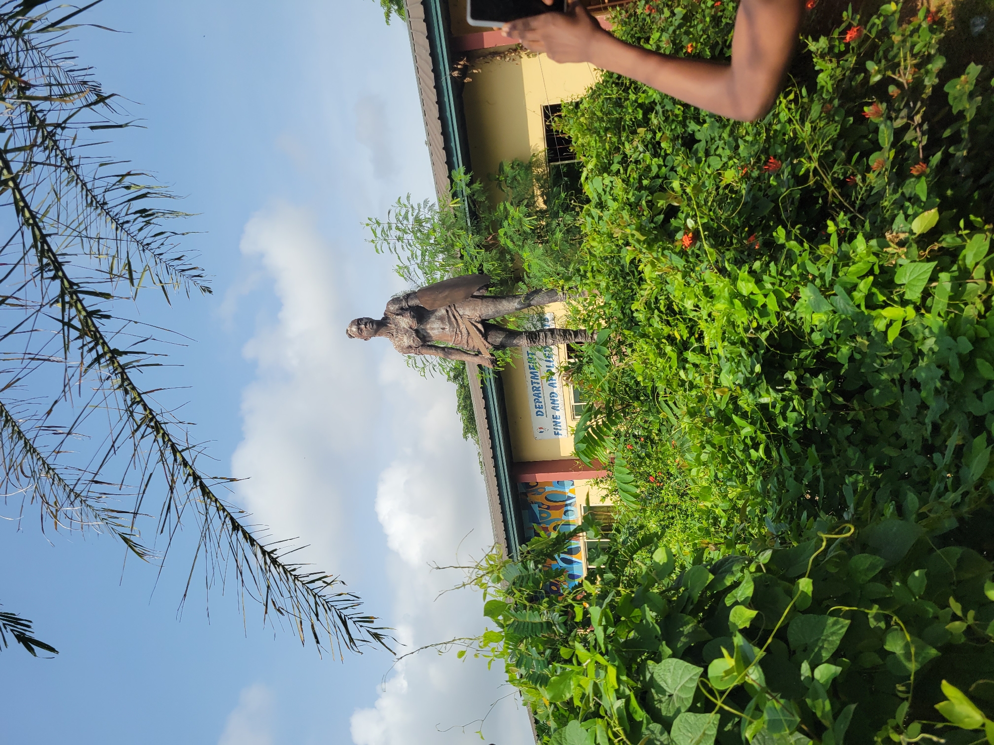 In an outdoor scene, a bronze statue of a woman in traditional attire stands prominently amidst lush greenery. The statue, appearing to be of African origin, is equipped with a shield and what looks like a spear. Behind the statue, a yellow building with a dark roof is visible, featuring the words "DEPARTMENT OF FINE AND APPLIED" on its facade. A person's arm and hand, holding a mobile phone, are captured in the foreground, suggesting the act of taking a photograph. The sky above is a clear blue with scattered white clouds, and palm fronds hang into the frame from the top left, adding to the tropical atmosphere. The overall setting suggests a location in a warm climate, possibly a cultural or educational institution.