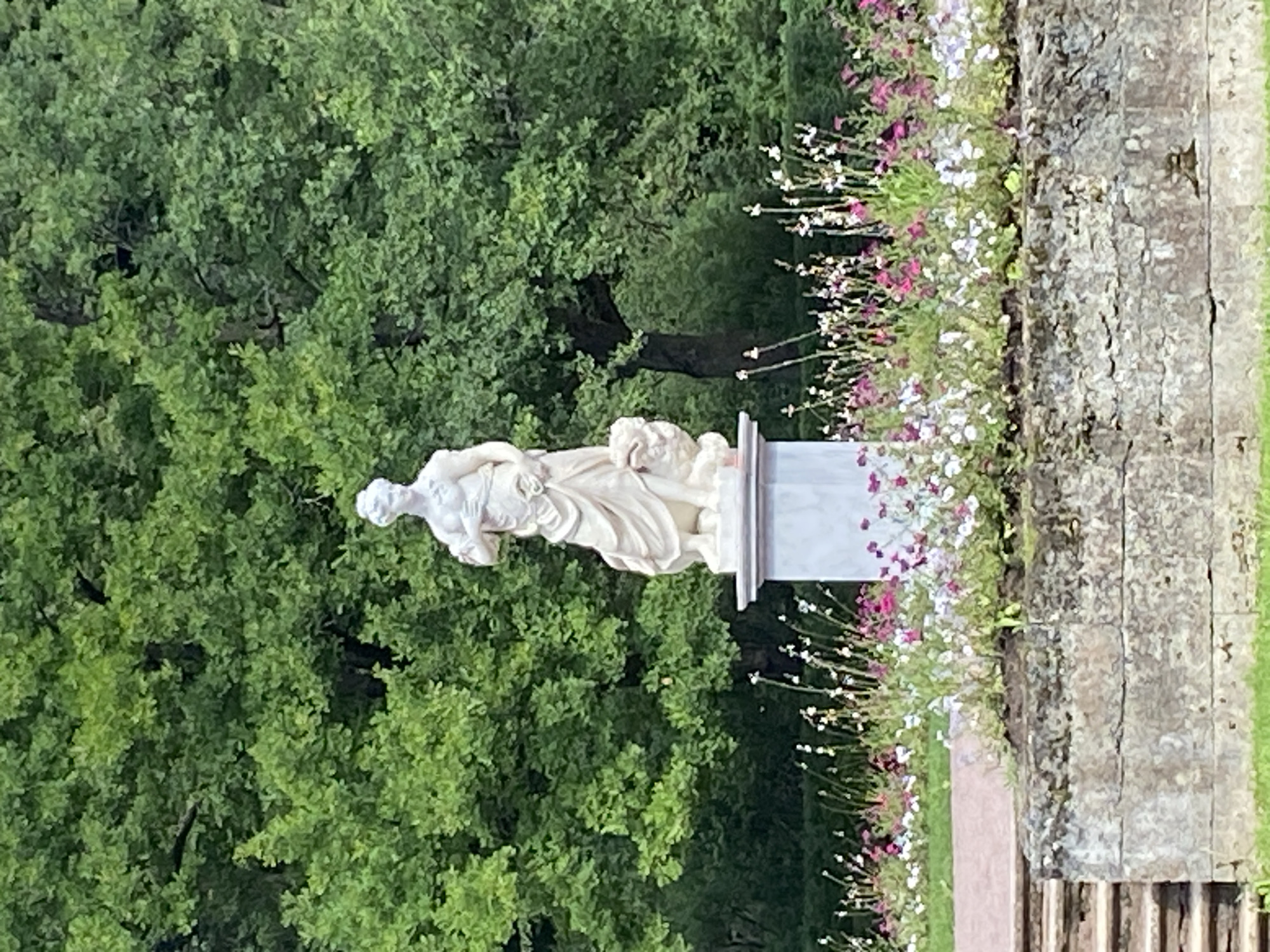 The image depicts a marble statue of a woman standing on a pedestal, situated in a garden setting in Pavlovsk, Russia. The statue is white and appears to be in classical style, with the woman draped in flowing robes and holding something in her hand. A lion or similar mythical creature is positioned at the base of the statue. The statue is surrounded by a vibrant flowerbed filled with pink, white, and purple blossoms. Behind the flowerbed, a stone wall with visible cracks and textures is present, with a set of stone steps leading up to it. The background consists of lush green trees, creating a natural and serene environment. The overall scene suggests a well-maintained garden or park, likely a historical or cultural site in Pavlovsk.