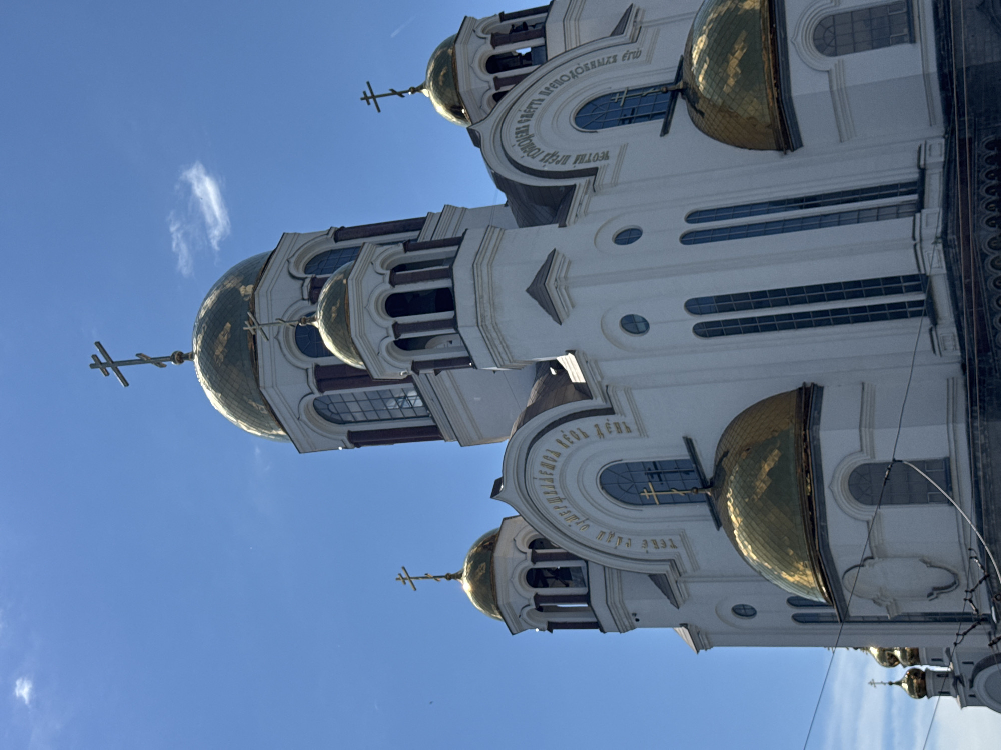 The image shows the Church on Blood in Yekaterinburg, Russia. The church is a white building with multiple golden domes and crosses. The sky is blue with a few wispy clouds. The camera angle is from below, looking up at the church. There are no people visible in the image. The church is a prominent landmark in Yekaterinburg, built on the site where Tsar Nicholas II and his family were executed.
