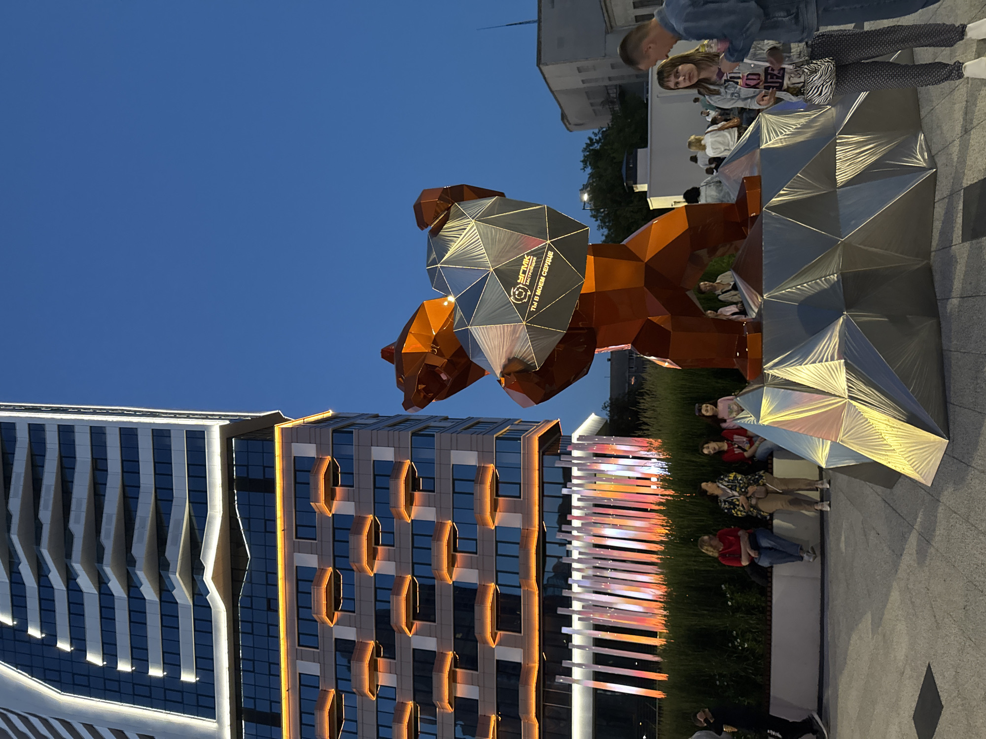 The image shows an outdoor art installation in Yekaterinburg, Russia, likely located in the city center. In the foreground, a large, geometric sculpture of a bear is prominently displayed. The bear is depicted in a rust-orange color and appears to be holding a faceted, silver object. The sculpture is situated on a similarly geometric, faceted base.
Behind the sculpture, a modern, multi-story building with distinctive horizontal bands of orange lighting is visible. The building has a sleek, contemporary design with numerous windows. In front of the building, a row of thin, illuminated vertical poles in varying shades of pink and orange creates a visually striking background.
Several people are present in the scene. A group of individuals is seated on a bench in the middle ground, some facing the sculpture and others conversing amongst themselves. Further back, other people can be seen walking and milling about. The sky is a clear, deep blue, indicating it is likely dusk or early evening. The overall impression is of a public space with modern architecture and art, designed for public enjoyment.