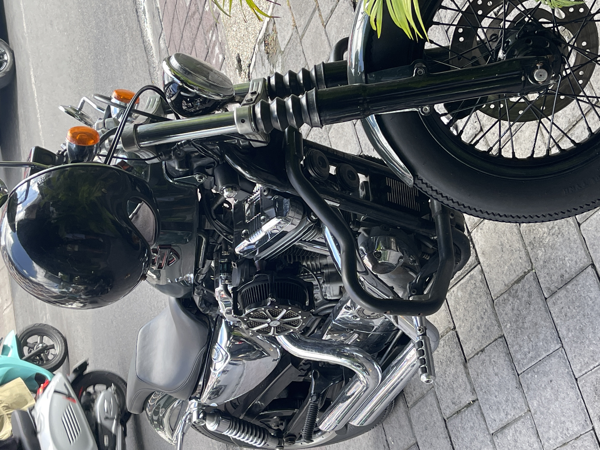 An eye-level medium shot captures a dark green Harley-Davidson motorcycle parked on a paved sidewalk in North Kuta, Indonesia. The motorcycle is the primary subject, with its chrome accents and black leather seat prominently displayed. A black helmet rests on the motorcycle's handlebars. In the background, a sliver of a teal scooter is visible. The lighting suggests it is daytime, and the overall atmosphere is that of a typical urban street scene.