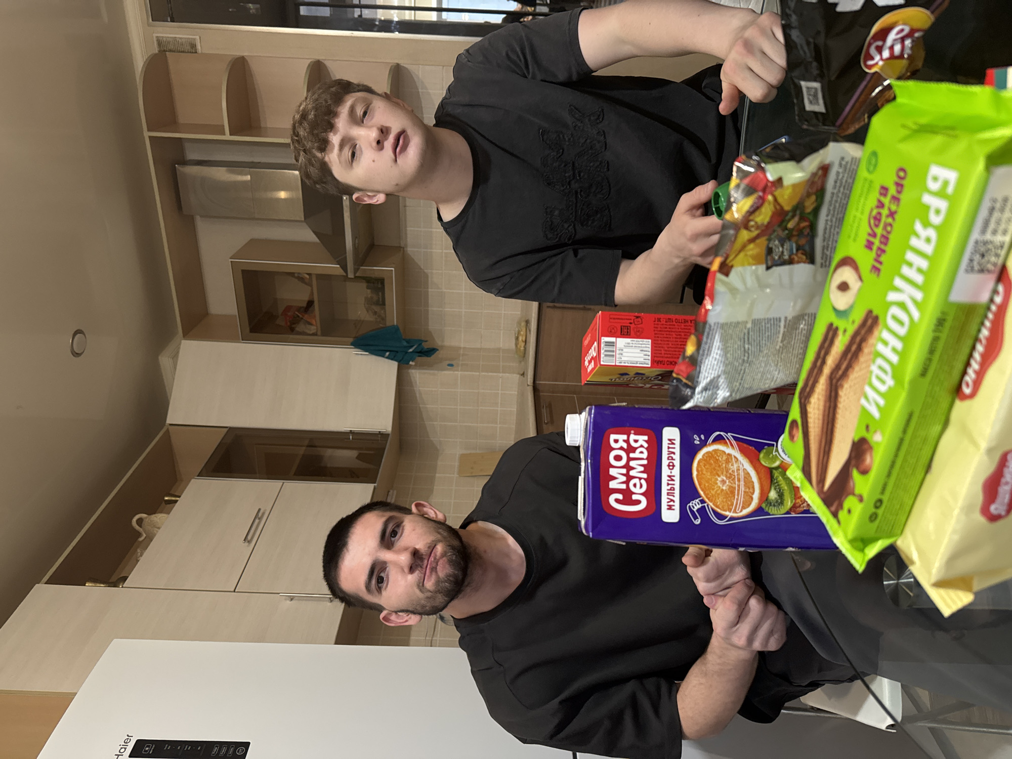 Two men are sitting at a table indoors, likely a kitchen given the cabinets, microwave, and refrigerator visible in the background. The refrigerator is a Haier brand. The men are both wearing black t-shirts. One man has shorter hair and a beard. The other man has curly brown hair. On the table are various packaged food items, including bags of potato chips (lays), a box of what appears to be candy, a carton of juice with text in Russian, and a package of waffles with a green wrapper that reads "БРЯНКОНФИ" and "ОРЕХОВЫЕ ВАФЛИ," also in Russian. The juice carton says "МОЯ Семья" and "МУЛЬТИ-ФРУТИ" in Russian. The location is likely in Russia, given the Russian text on the packaging. There is no information to specify that this is in the city of Kazan. The men appear to be preparing to eat or are in the process of opening the various packages.