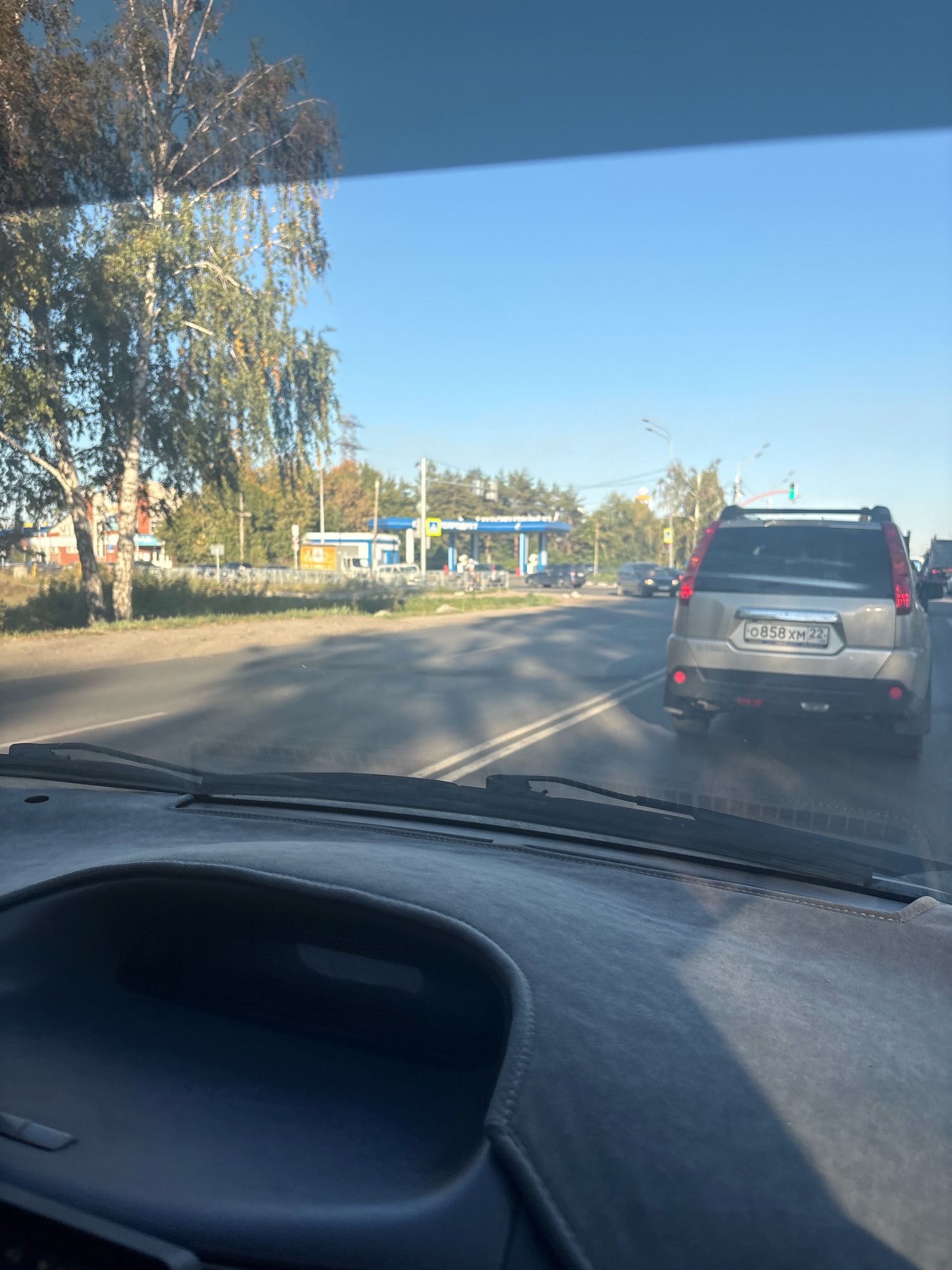 This is a photograph taken from inside a vehicle in Barnaul, Russia. The camera is positioned behind the dashboard, looking through the windshield. The foreground shows the interior of the car, specifically the textured dashboard.
In the middle ground, a silver SUV with license plate "0858 XM 22" is visible in the right lane of a paved road. The SUV has its brake lights illuminated. Other vehicles are present in the distance, including a black car in the left lane and a white truck at a gas station on the left side of the road. The gas station has blue and white awnings and appears to be operational.
The background features a clear blue sky and a line of trees, some of which are displaying autumn colors. The overall environment suggests a sunny day. The scene is consistent with being on a main road within a city or its outskirts.