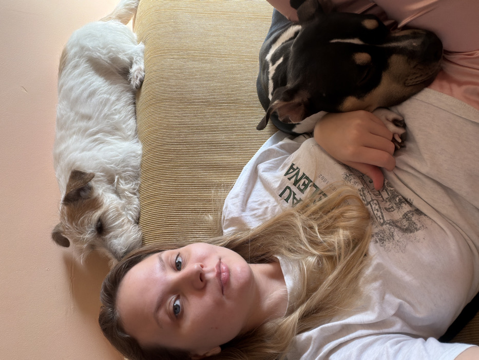The image depicts an indoor scene with one human and two dogs. A fair-skinned female, appearing to be in her twenties or thirties, with long, light brown hair, is positioned in the foreground, facing the camera directly with a neutral expression. She is wearing a light grey short-sleeved t-shirt with a dark graphic print featuring text and an architectural design, and light pink or peach-colored pants. Her left hand rests on the body of a dog sleeping on her lap.
Two dogs are present. One, a white and tan wire-haired terrier type, is lying prone on the top edge of a mustard-yellow, vertically ribbed fabric couch backrest, with its head facing left and eyes appearing closed. The second dog, a black and tan short-haired breed, possibly a mixed breed with markings similar to a miniature pinscher or a similar terrier mix, is resting on the female's lap/upper torso, with its head to the right and eyes closed. Its ear is visible, and it appears to be either sleeping or deeply relaxed.
The background shows a wall painted in a light peach or salmon color. The exact location within Tel-Aviv, Israel, cannot be determined from the visual information provided in the image.