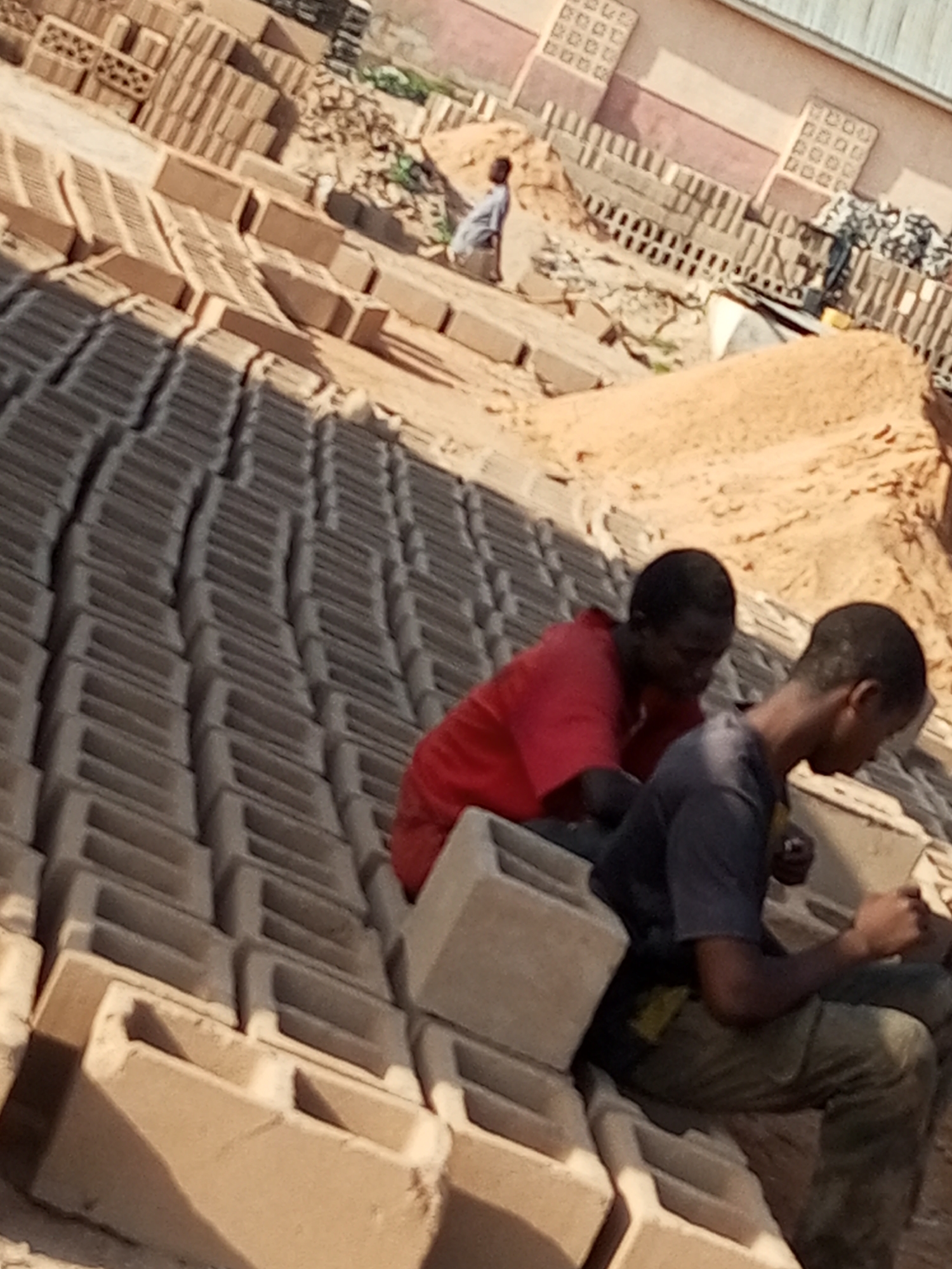 A low-angle, outdoor, medium shot shows two men sitting on concrete blocks arranged in rows. The man on the left, wearing a red shirt, is partially obscured by the man on the right. The man on the right, wearing a dark blue t-shirt and green pants, is looking down at something in his hands. They appear to be in a construction or material yard, possibly in Tempure, Nigeria, given the context. The background consists of more stacked blocks, piles of sand, and a building with a light-colored facade and a metal roof. The lighting suggests it is daytime, possibly late morning or afternoon, with the sun casting shadows. The scene is gritty and industrial, with many unfinished building materials present.