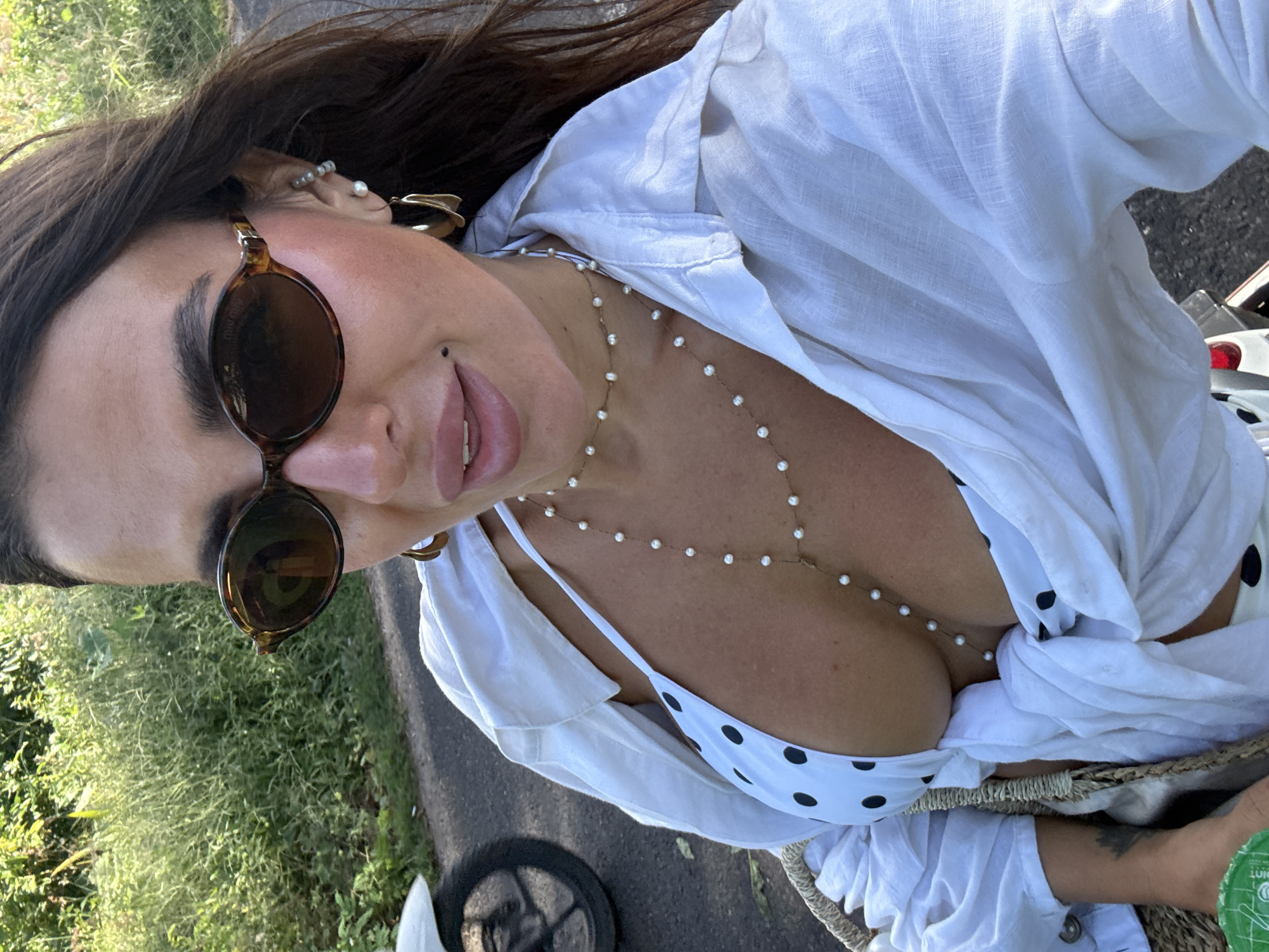 A fair-skinned woman with brown hair and sunglasses takes a selfie outside in Kuta Selatan, Indonesia. She wears a white blouse open to show a white bikini top with black polka dots, and a long pearl necklace. The background includes green foliage and pavement, suggesting she is on a road or pathway. A scooter wheel is visible, partially obscured by the background. She is holding a bag with a green container inside.