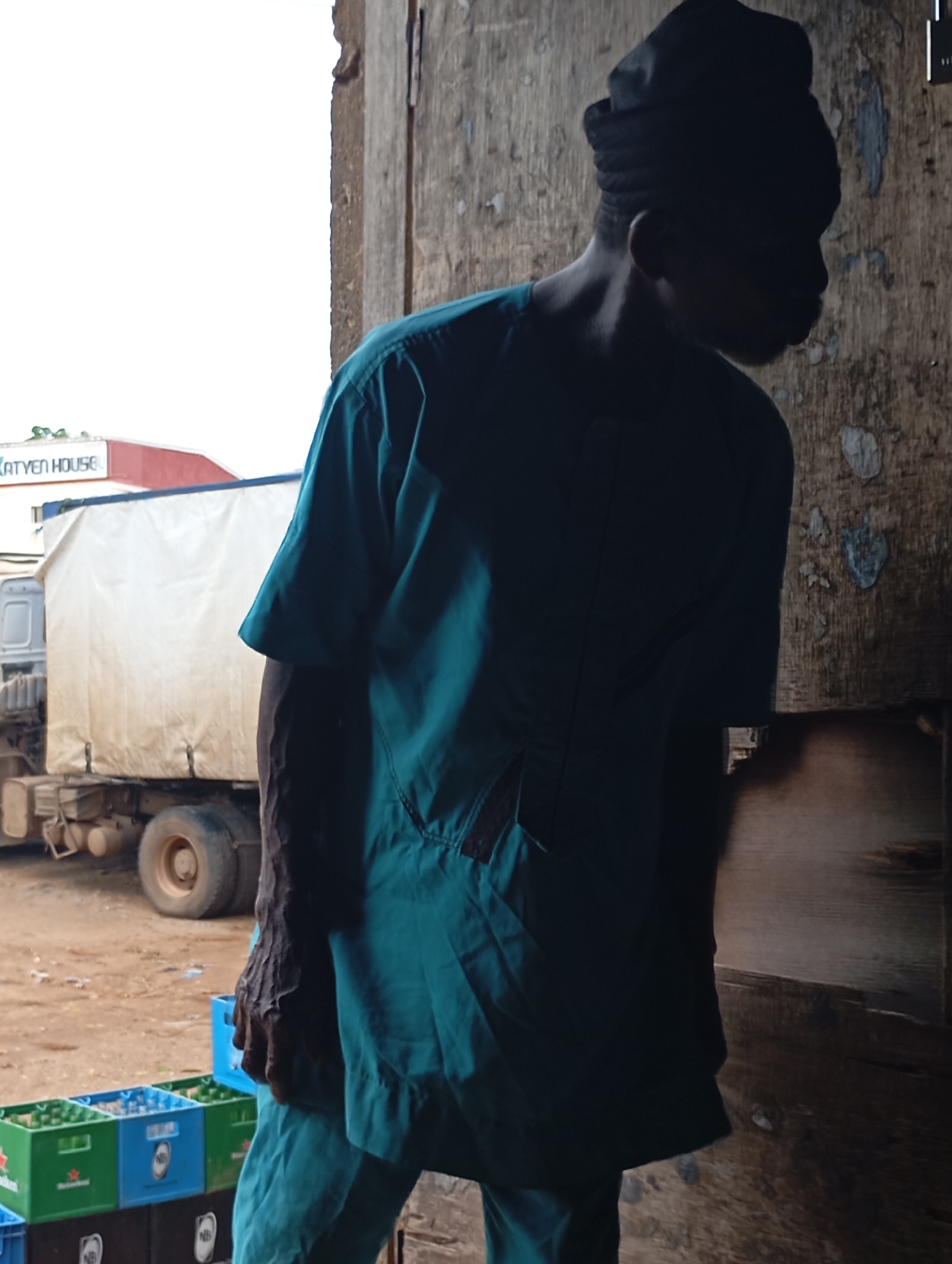 In Shendam, Nigeria, a man wearing a dark turban and a teal uniform stands partially silhouetted against a rough wooden wall. He is positioned to the right of the frame, with his back to the viewer. To his left, a large, white-covered truck is parked on a dusty lot. In the foreground, stacked crates of green and blue bottles, some with Heineken logos, are visible. A red and white building with the text "KATYEN HOUSE" is partially visible in the background behind the truck. The overall scene suggests a delivery or storage area.