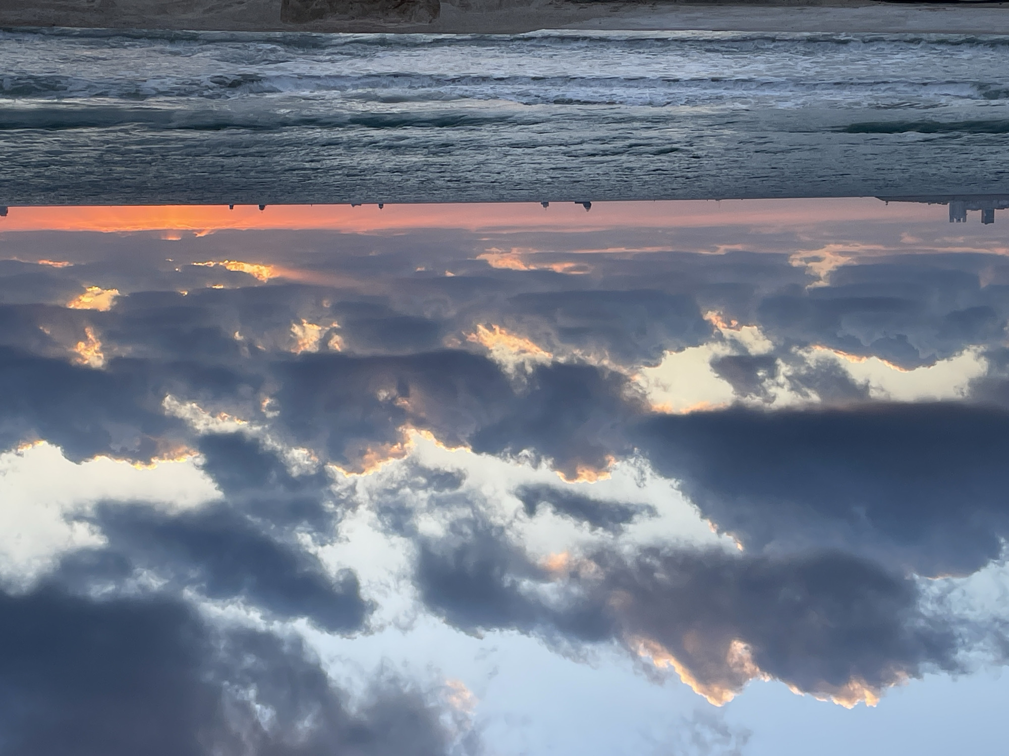 This is a vertical, wide-angle photograph of a sunset over the Mediterranean Sea, viewed from a beach in Haifa, Israel. The image is oriented with the sea and sky at the top and bottom respectively, and the beach at the very top edge.
The foreground, at the top of the image, is a sandy beach, with some visible texture suggesting footprints or ripples. Below the beach is the sea, characterized by a foamy, wave-swept surface and a range of blues and grays.
The horizon line is dominated by a vibrant sunset. A band of bright orange and pink light stretches across the sky, indicating the setting sun. Silhouetted against this light, along the distant horizon, are the faint outlines of buildings, suggesting a coastal city.
Above the sunset, the sky is filled with dramatic clouds. These clouds are a mix of dark grays and lighter whites and blues, with edges illuminated by the setting sun, creating a dramatic and textured sky. The light filtering through the clouds gives them a warm, golden glow in places.
The overall mood is serene and awe-inspiring, capturing the beauty of a coastal sunset. The time of day is clearly sunset, and the location is implied to be a beach in Haifa, Israel due to the presence of the Mediterranean Sea and the distant city skyline. No people or specific objects are clearly identifiable, and there is no visible text in the image.
