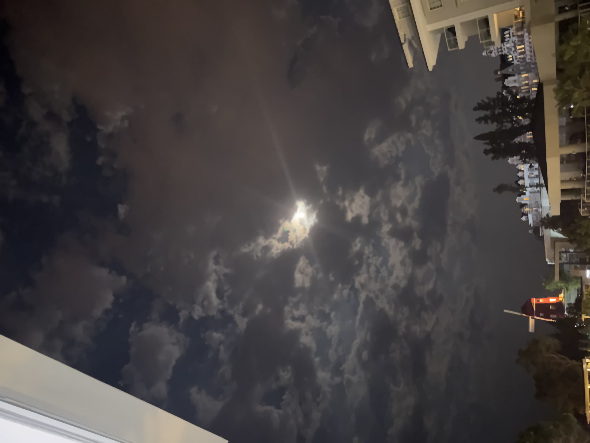 The image shows a nighttime view of the city of Kemer, Turkey. In the foreground, there is a white building with balconies. In the middle ground, there are several ornate buildings illuminated with lights, and a red windmill. In the background, the sky is dark and cloudy with a bright moon shining through. The scene appears to be a resort or hotel area.
