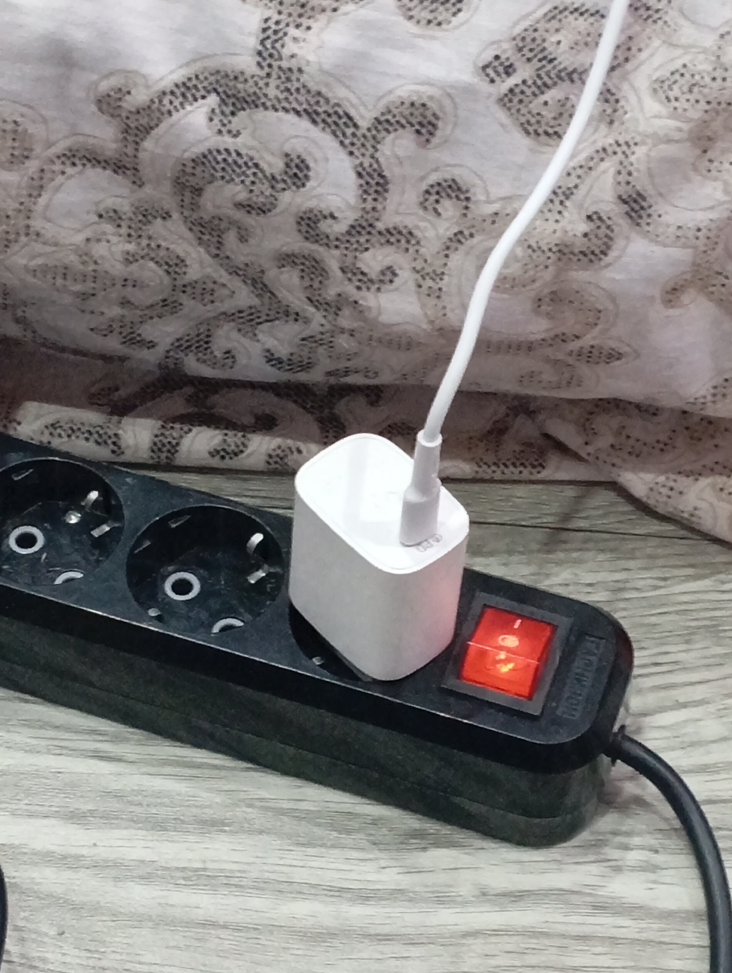 The image captures an indoor scene, likely in a home setting, with an emphasis on electronic accessories. In the foreground, a black power strip with three visible Schuko-type sockets rests on a light-colored wooden surface, possibly a floor or table. A white, rectangular charging adapter is plugged into the rightmost socket of the power strip. A white charging cable extends upwards from the adapter, presumably connecting to a device that is out of frame. The power strip features a red illuminated rocker switch, indicating it is currently powered on. Faint text "PD" is visible on the side of the white charging adapter, suggesting it supports Power Delivery. The brand name "FADUYA" is faintly visible on the power strip near the switch. The background consists of a crumpled or draped textile with a repeating, ornate pattern in shades of grey and white, providing a soft, textured backdrop. The overall impression is one of a routine charging activity taking place. The type of power sockets is consistent with the provided location context of Krasnodar, Russia.