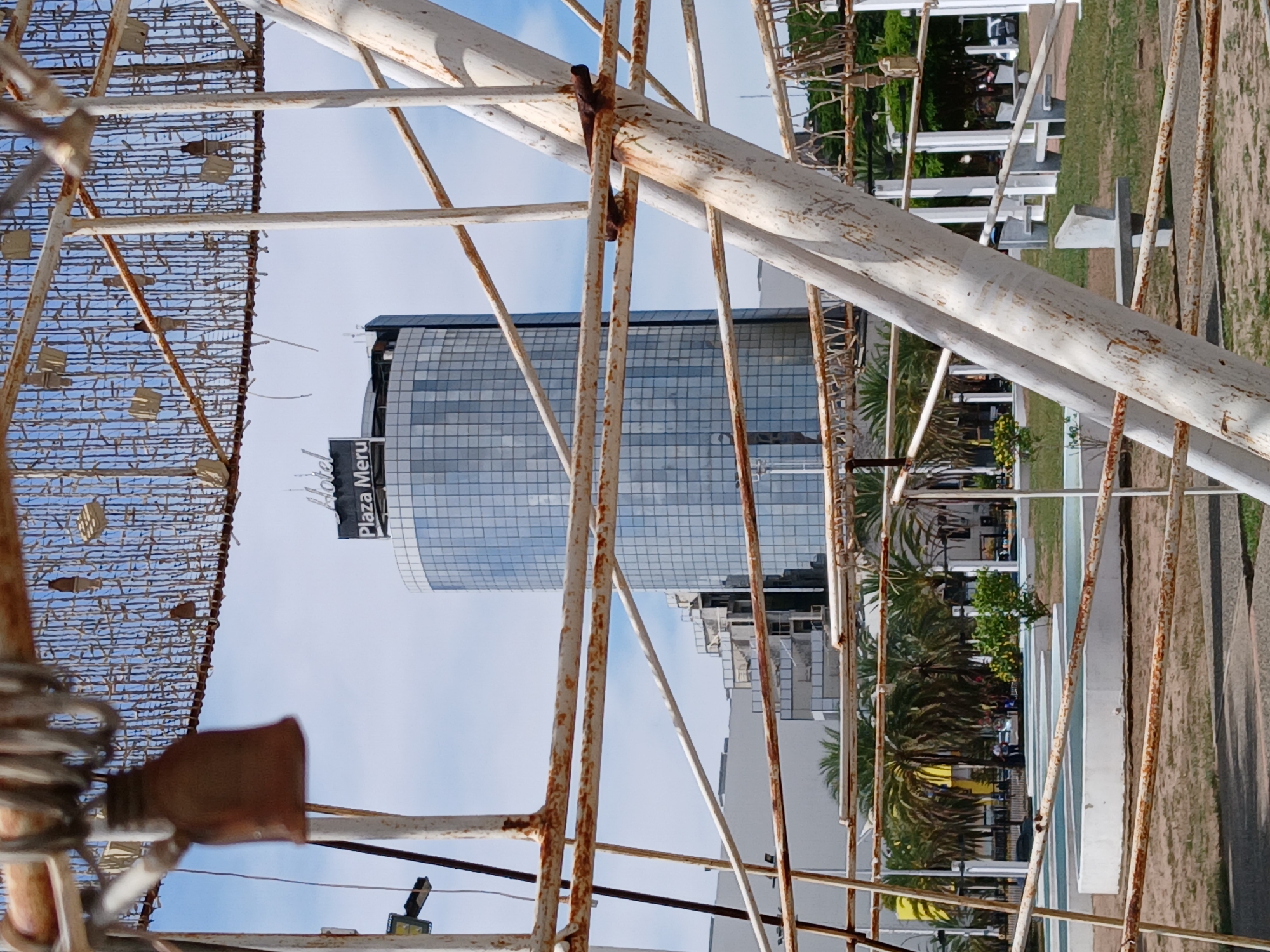 The image depicts a view of the Plaza Meru office building in Ciudad Guayana, Venezuela, as seen through a weathered metal structure. The building is a modern, curved glass skyscraper. The foreground structure appears to be an old, possibly decorative piece of infrastructure, showing signs of rust and disrepair. The environment includes landscaping with trees and a pedestrian walkway.