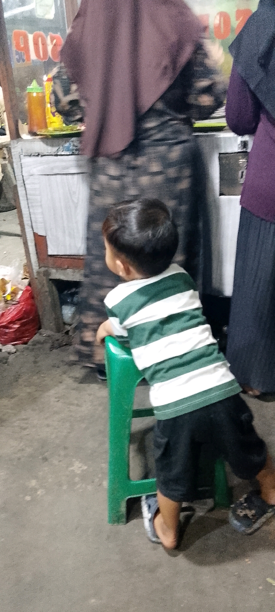 The image shows a young child from behind, wearing a green and white striped shirt and black shorts, leaning on a green plastic stool. The child's feet are bare in one sandal and the other is on the floor. In the background, two women are visible, one wearing a purple headscarf and patterned dress, and another wearing a dark purple headscarf and long black skirt. Behind them is a stall with what appears to be food items and bottles on a counter. The scene appears to be outdoors, possibly on a street, with a concrete floor. The setting is identified as Satriamekar, Indonesia.