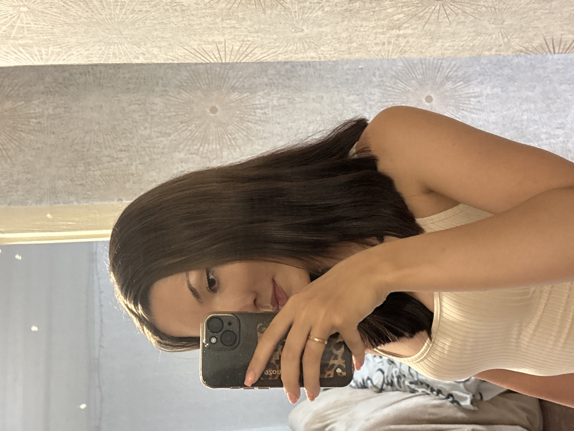 The media file shows a young woman taking a selfie in a bedroom. She has long, dark hair and is wearing a white tank top. She is holding a smartphone with a leopard print case and a gold ring is visible on her finger. The background shows a textured wallpaper with a sunburst pattern and a slightly blurred bed with a patterned duvet. The lighting suggests it is daytime, likely indoors. The woman's expression is neutral, and the overall mood is casual. No visible text is present in the image. The location context is Inskoy, Russia.