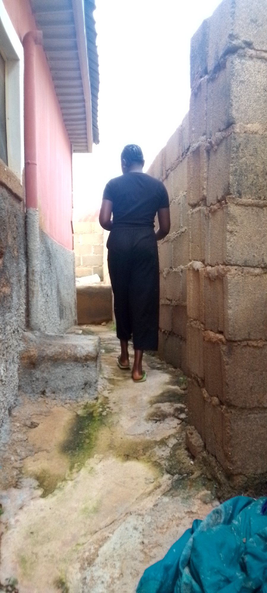 A person walks away from the camera down a narrow, outdoor alleyway. The person is wearing a black t-shirt and black pants, and green flip-flops. The alley is bordered on the left by a pink building with a corrugated metal roof overhang, and on the right by a wall constructed of concrete blocks. The ground of the alley appears to be dirt and concrete, with some patches of green algae or moss. A teal cloth is visible on the ground in the lower right portion of the image. The sky above is bright and overcast.