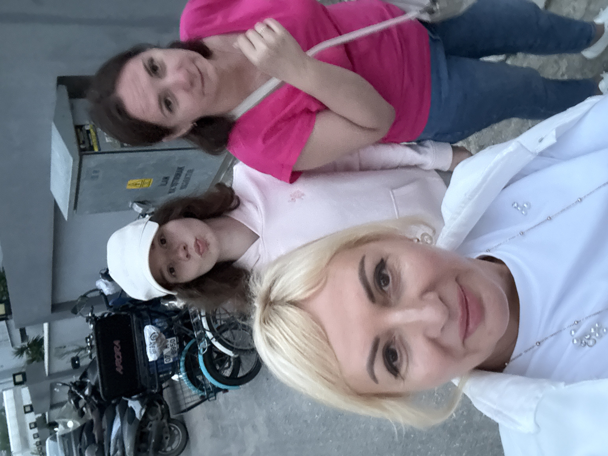 Three women are posing for a selfie outdoors. The woman in the foreground has blonde hair and is wearing a white shirt and jacket, along with a silver necklace and pearl earrings. Behind her to the left is a younger woman with brown hair and a white cap, wearing a light pink shirt. In the top of the frame is a woman with dark hair, wearing a pink shirt and blue jeans, carrying a white purse. Behind the women is a utility box and parked motorbikes with a box reading "Arora". The scene appears to be in an urban area.