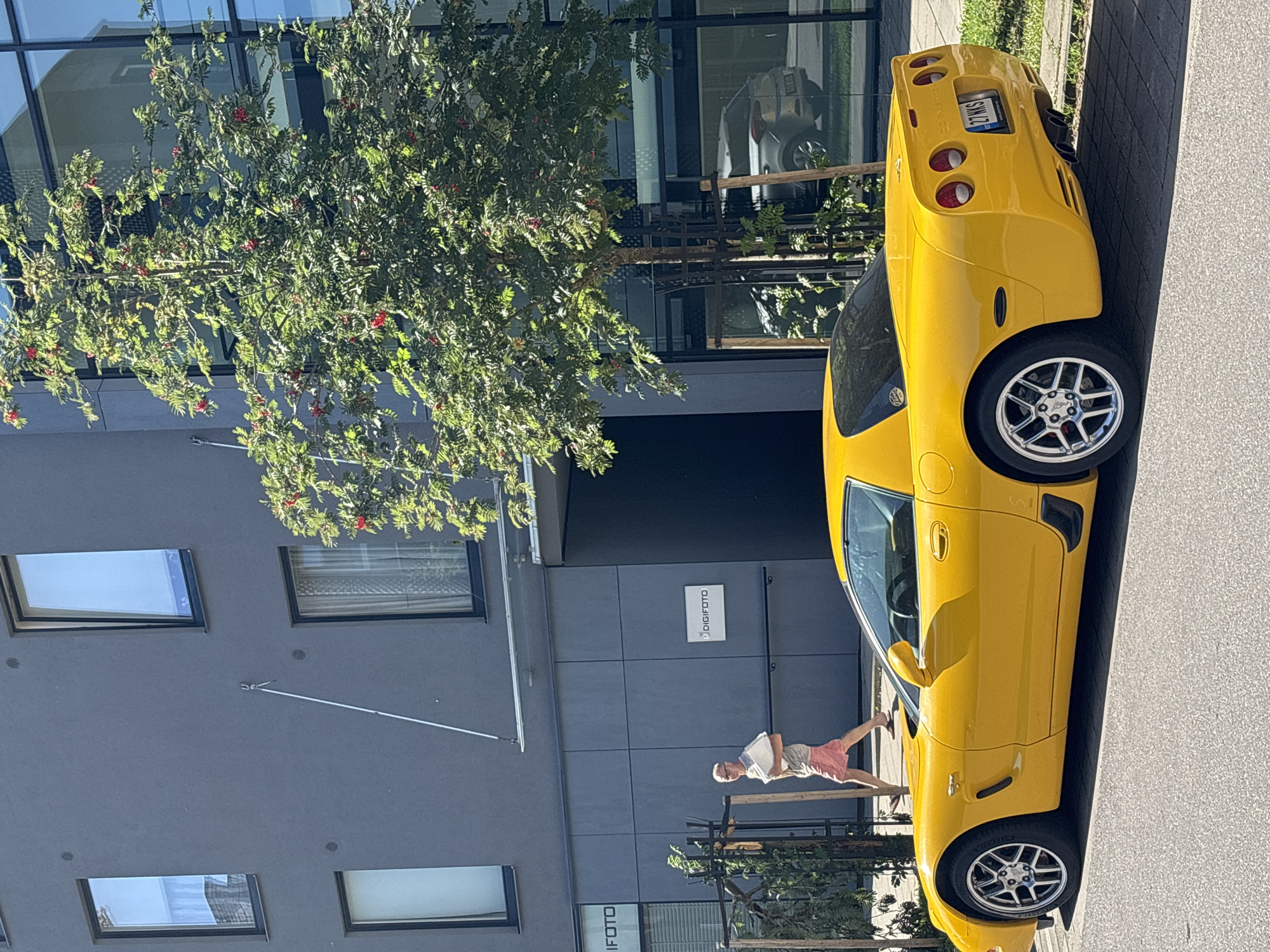 An eye-level, wide shot captures a bright yellow Chevrolet Corvette C5 parked curbside on a paved street in front of a modern, grey building in Tallinn, Estonia. The building features large, dark-framed windows, some reflecting the sky, and a prominent sign for "DIGIFOTO" is visible on the facade. A tall, leafy tree with red berries stands to the right of the car, obscuring part of the building's windows and reflecting another vehicle. A man, wearing a light t-shirt and pink shorts, is walking away from the camera on the sidewalk in front of the building, carrying a stack of white papers. The car's license plate reads "27 WKS". The scene is illuminated by bright sunlight, casting shadows on the street and sidewalk.