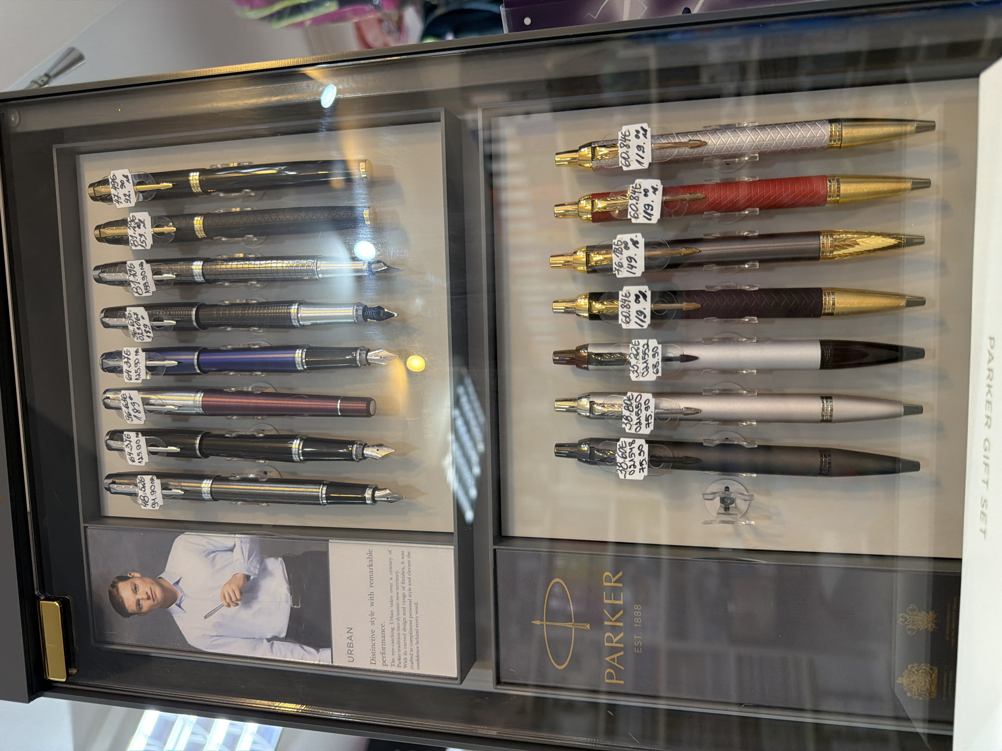 The image shows a display case of Parker pens. On the left side are Parker Urban pens with a picture of a man holding one. The right side displays Parker gift sets. Each pen has a price tag in euros and leva. The sign says Parker was established in 1888. While the location is Plovdiv, Bulgaria, it cannot be determined whether the image was taken inside a store or elsewhere in the city.