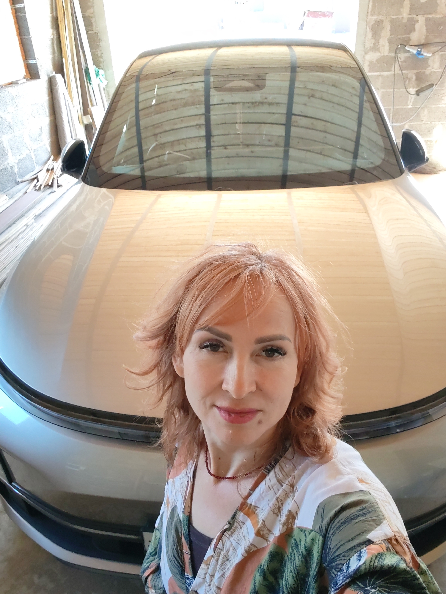 A woman is standing in front of a silver car in what appears to be a garage in Старое Михайловское, Russia. The garage walls are constructed of cinder blocks, and there are various pieces of lumber stacked against one wall. The car is the primary subject, and the woman is positioned slightly to the right of the frame. She is wearing a patterned top and a red necklace, and she is looking directly at the camera. The reflections in the car windshield suggest that the garage has a corrugated metal roof or door.