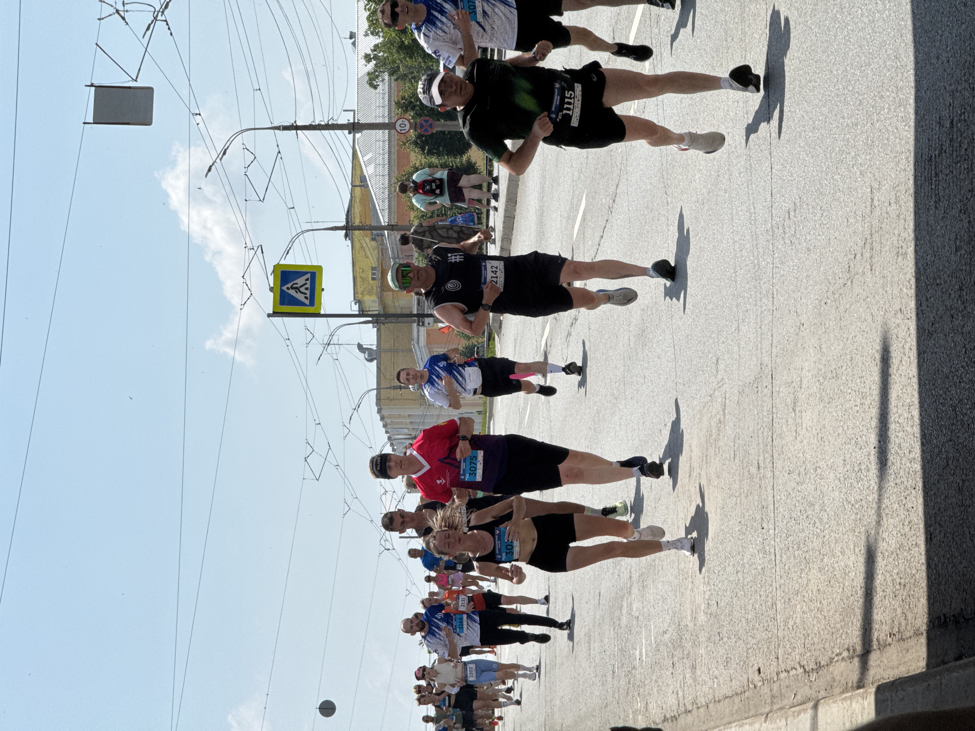 The image depicts a marathon race taking place on a street in Izhevsk, Russia, likely on a sunny day given the clear blue sky and shadows cast by the runners and overhead wires. In the foreground, several runners are actively participating in the race, their faces showing exertion. A female runner in a black sports bra and shorts is prominent, her arms pumped as she runs. To her right, a male runner in a black tank top and shorts, wearing a headband and sunglasses, is also visible. Further to the right, another male runner in a black t-shirt and shorts, wearing a baseball cap, is nearing the front. Behind them, a larger group of runners continues down the street, some wearing bib numbers. The street itself is paved and marked with white lines. Overhead, electrical wires and traffic lights suggest an urban environment. In the background, buildings and trees can be seen, consistent with a city setting. A pedestrian crossing sign is also visible, further confirming it's a public road within the city.
