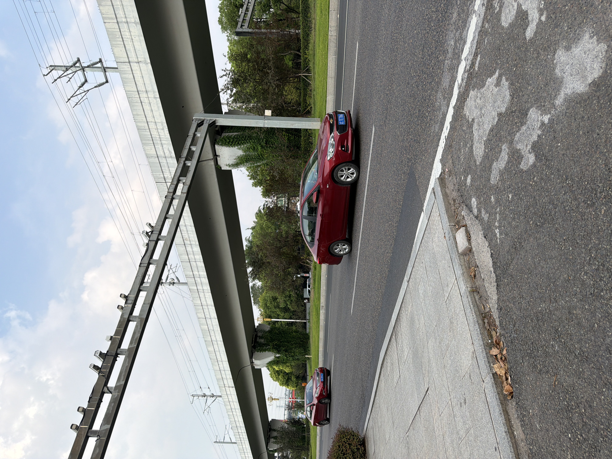 The image shows a street scene in Jiangjin District, Chongqing, China, with a prominent overpass structure. The overpass, likely a highway or elevated railway, has a grey concrete underside supported by large pillars. Attached to the underside of the overpass are numerous surveillance cameras, facing downwards and outwards. Several electrical wires and power line poles are visible above the overpass.
On the road beneath the overpass, two red sedans are driving. The car in the foreground is closer to the camera and is in the right lane, while another red sedan is further down the road in the left lane. The road surface is asphalt with white lane markings. To the left of the road, there is a paved sidewalk with square grey tiles and a concrete curb. Dry leaves are scattered along the edge of the sidewalk and curb. Greenery, including trees and bushes, is visible in the background on both sides of the road. The sky above is partly cloudy with a mix of white clouds and blue sky. The overall impression is an urban environment with infrastructure for transportation and surveillance.